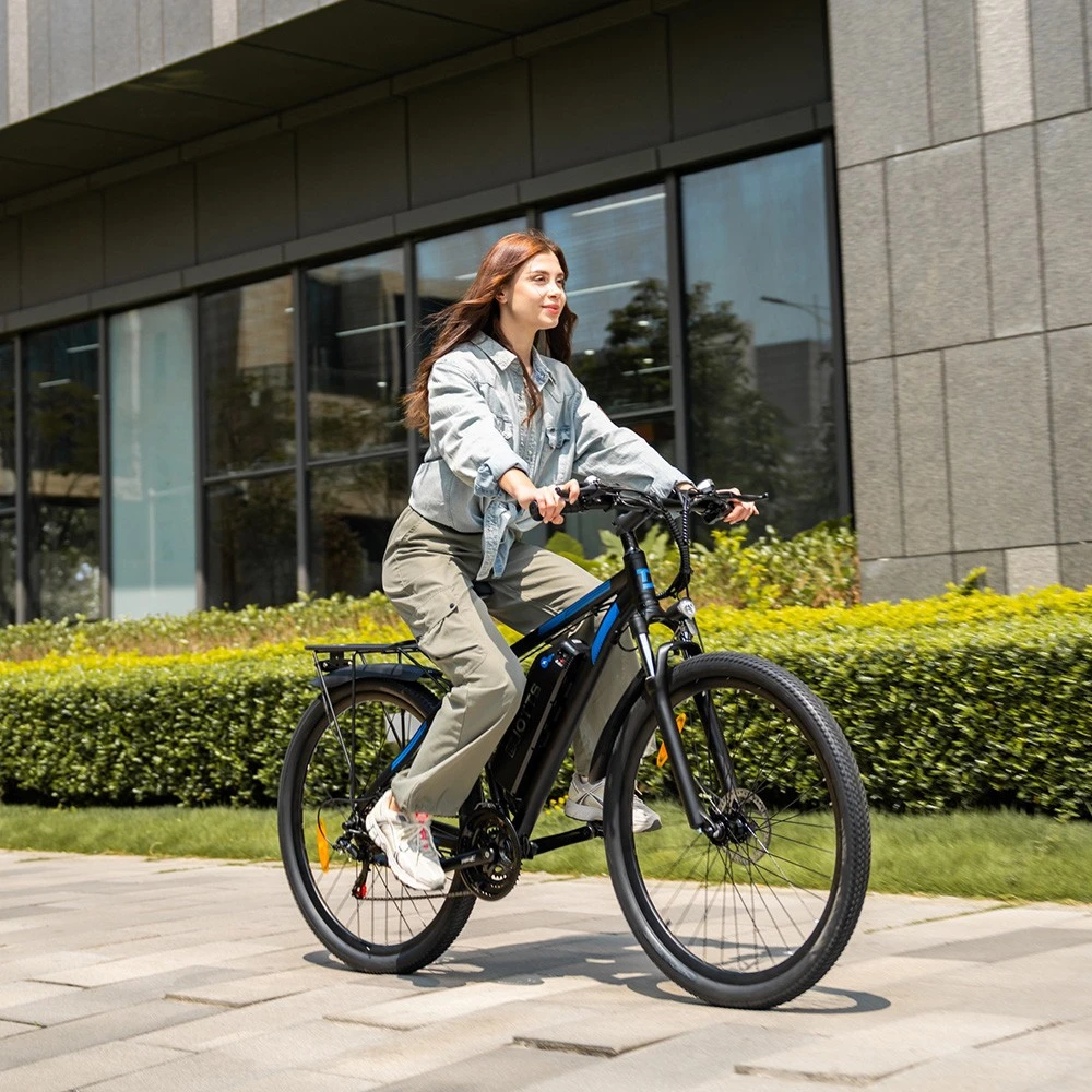 Una mujer monta una bicicleta eléctrica negra por un camino pavimentado, pasando arbustos y un edificio moderno.