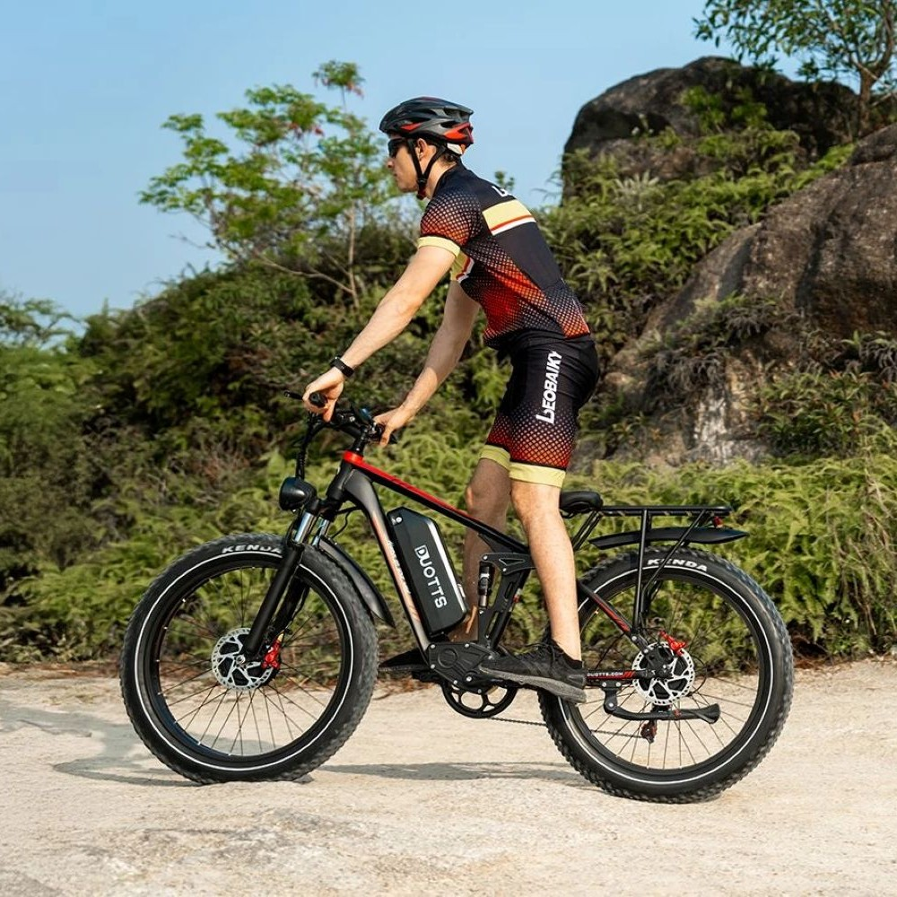 Hombre con casco y ropa de ciclismo monta una e-bike en camino de tierra, arbustos verdes detrás.