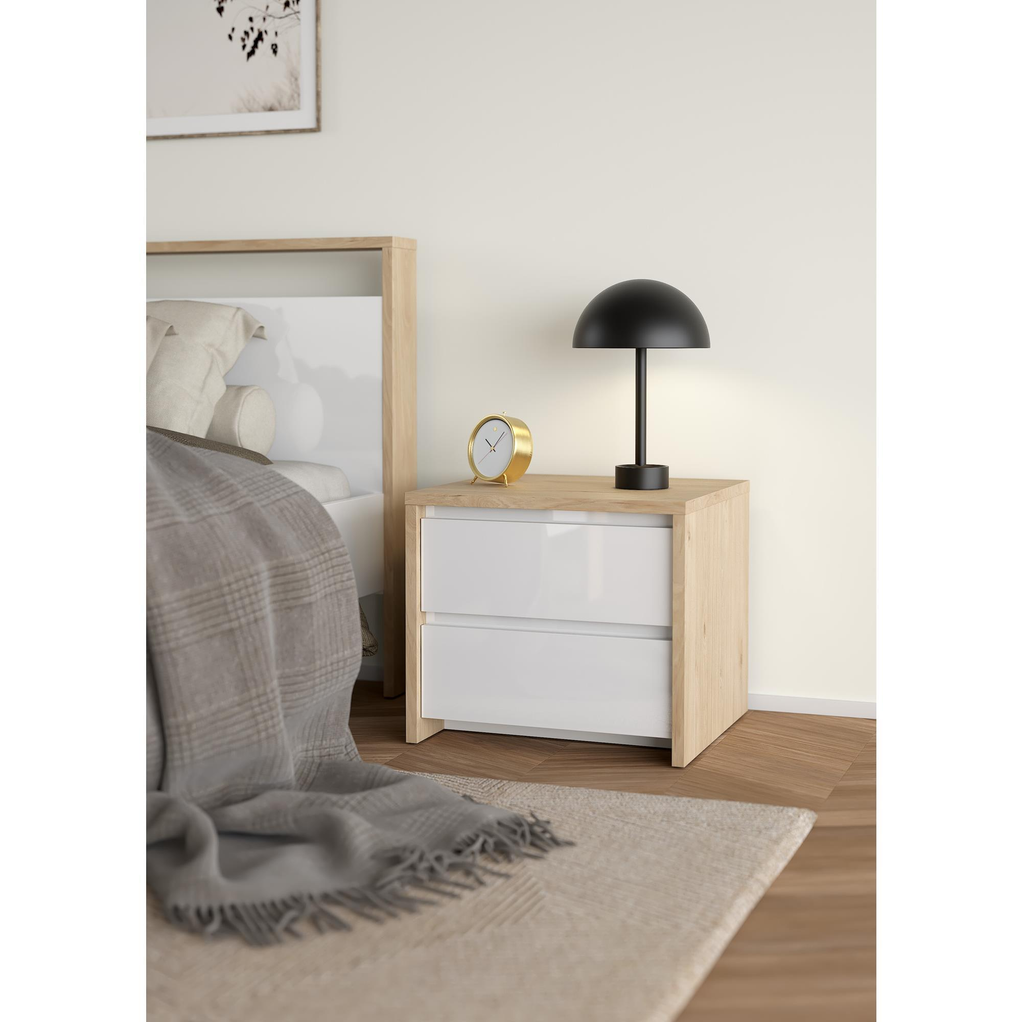 Una mesita de noche de madera con cajones blancos, lámpara, reloj y cama al fondo.