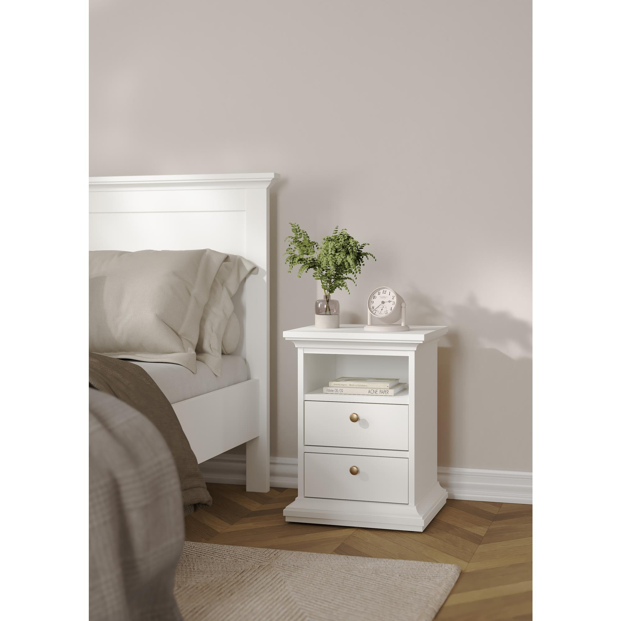 Una mesita de noche blanca junto a una cama, con reloj y planta.