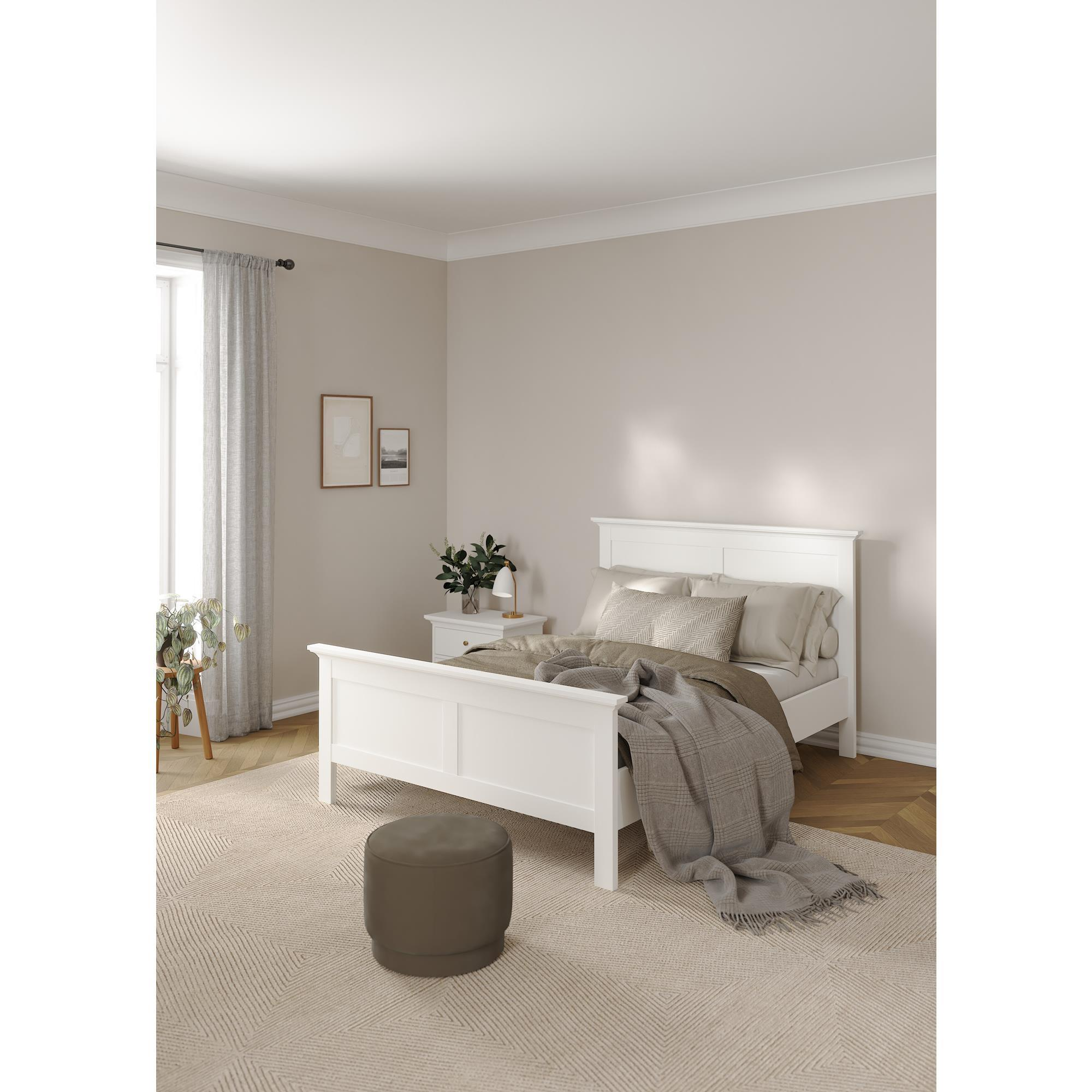Una cama blanca con cojines y manta, mesita de noche con lámpara.