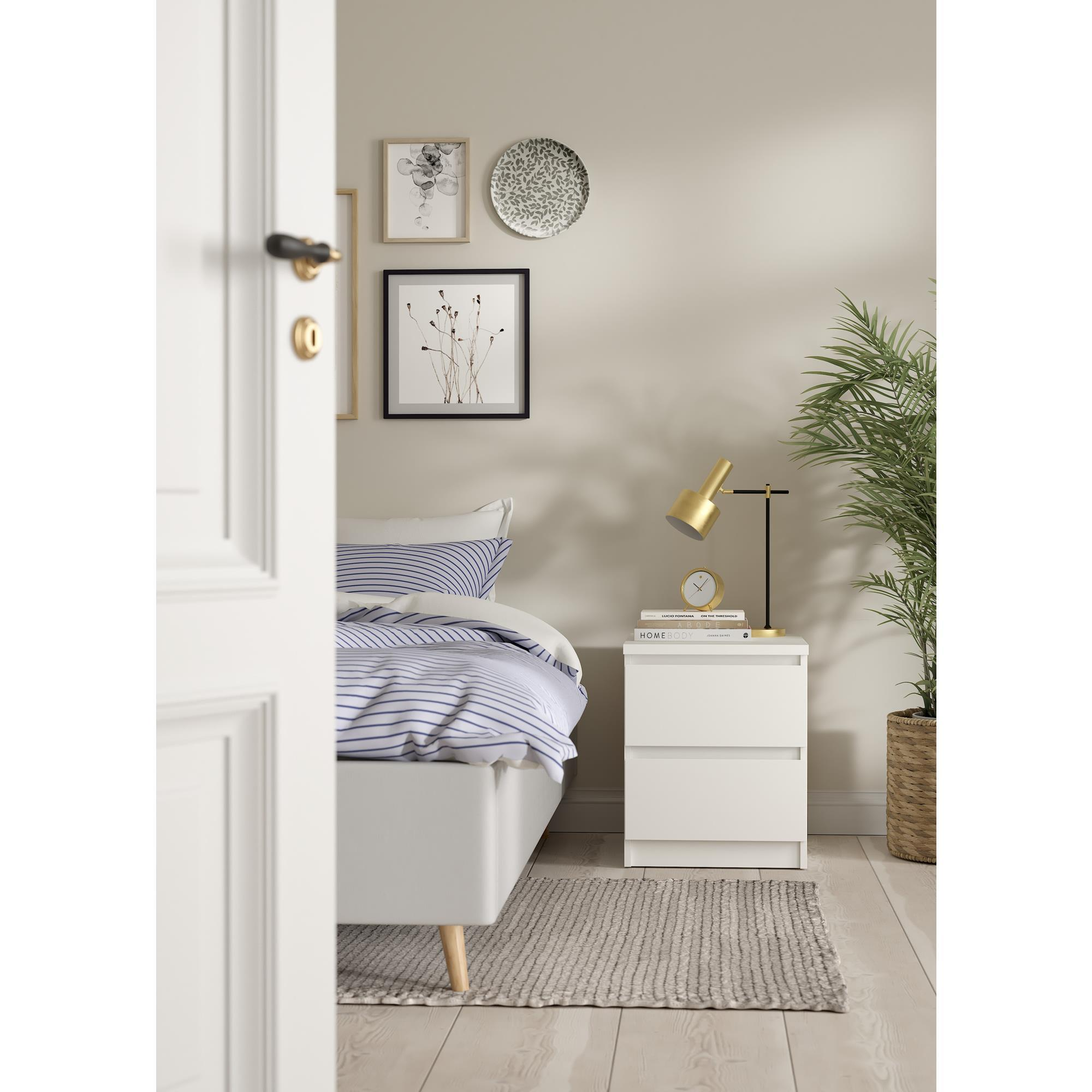 Un dormitorio con cama blanca, mesita de noche, lámpara y plantas.