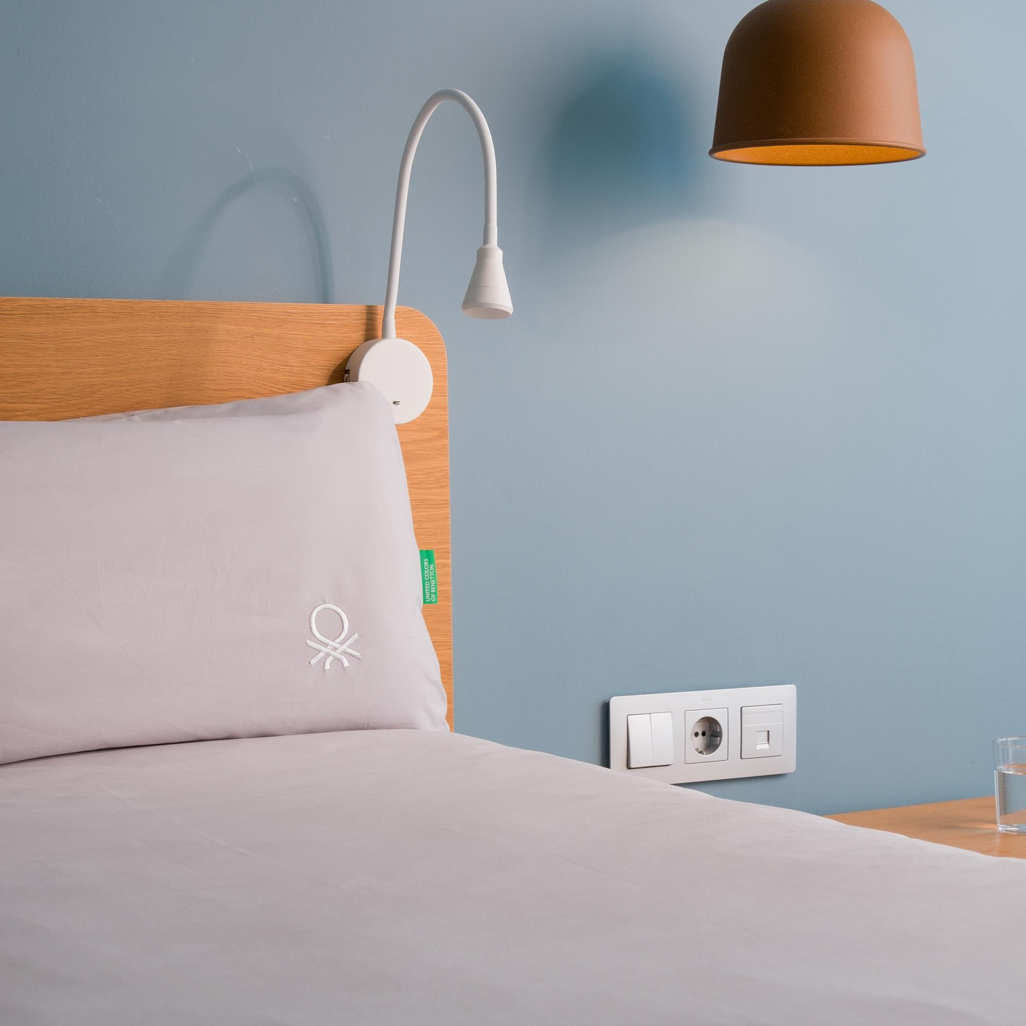 Una cama con almohada y edredón gris claro, lámpara de pared y enchufe.