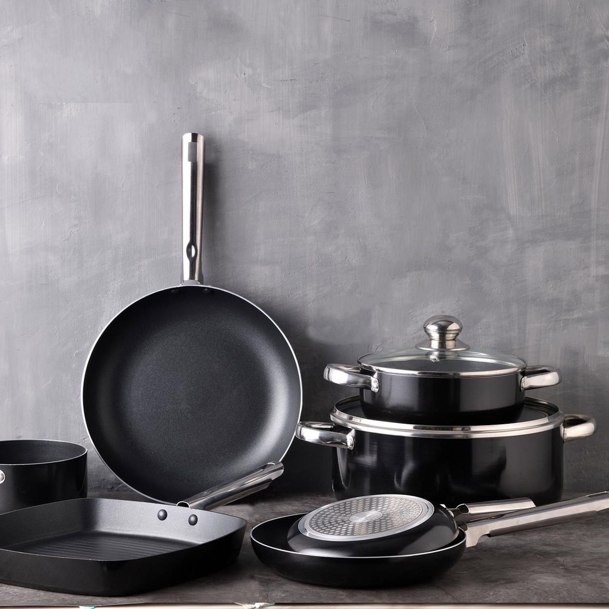 Varios utensilios de cocina negros, incluyendo sartenes y ollas, se muestran sobre fondo gris.