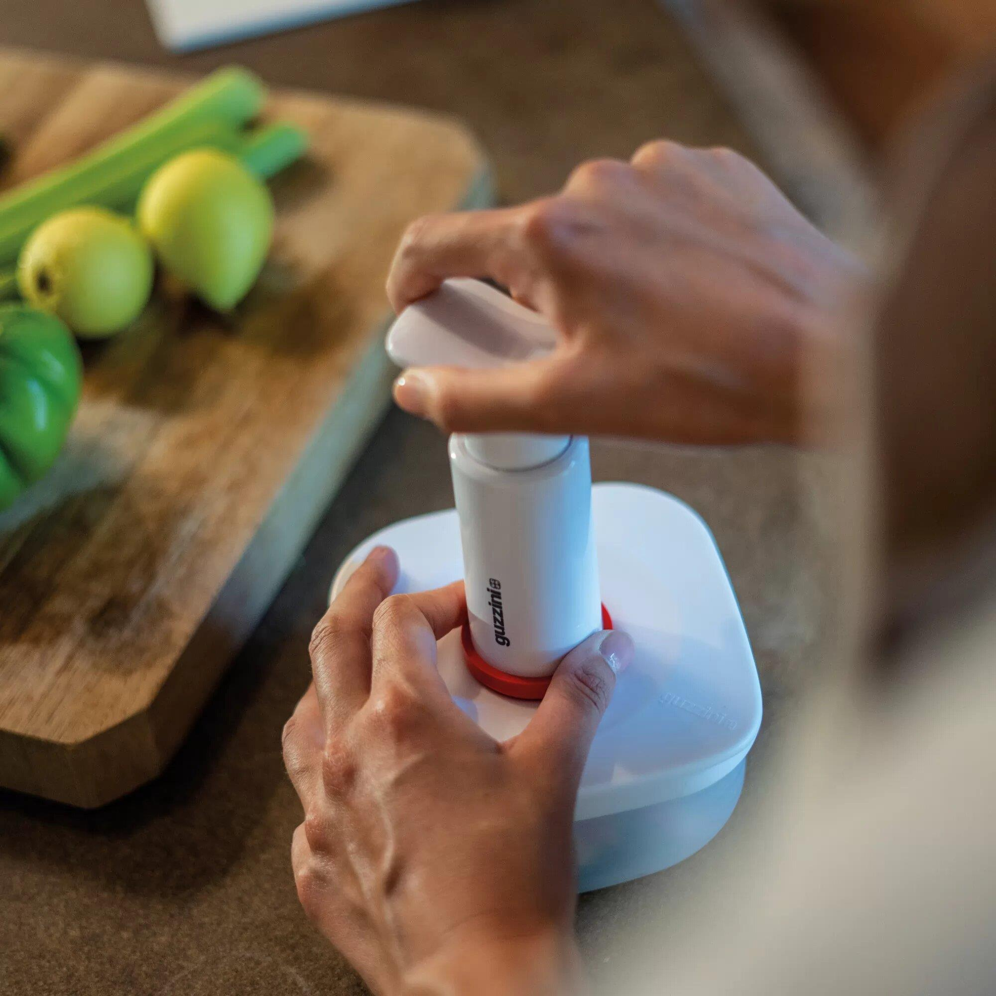Manos operan un sellador al vacío blanco con anillo rojo junto a verduras en tabla.