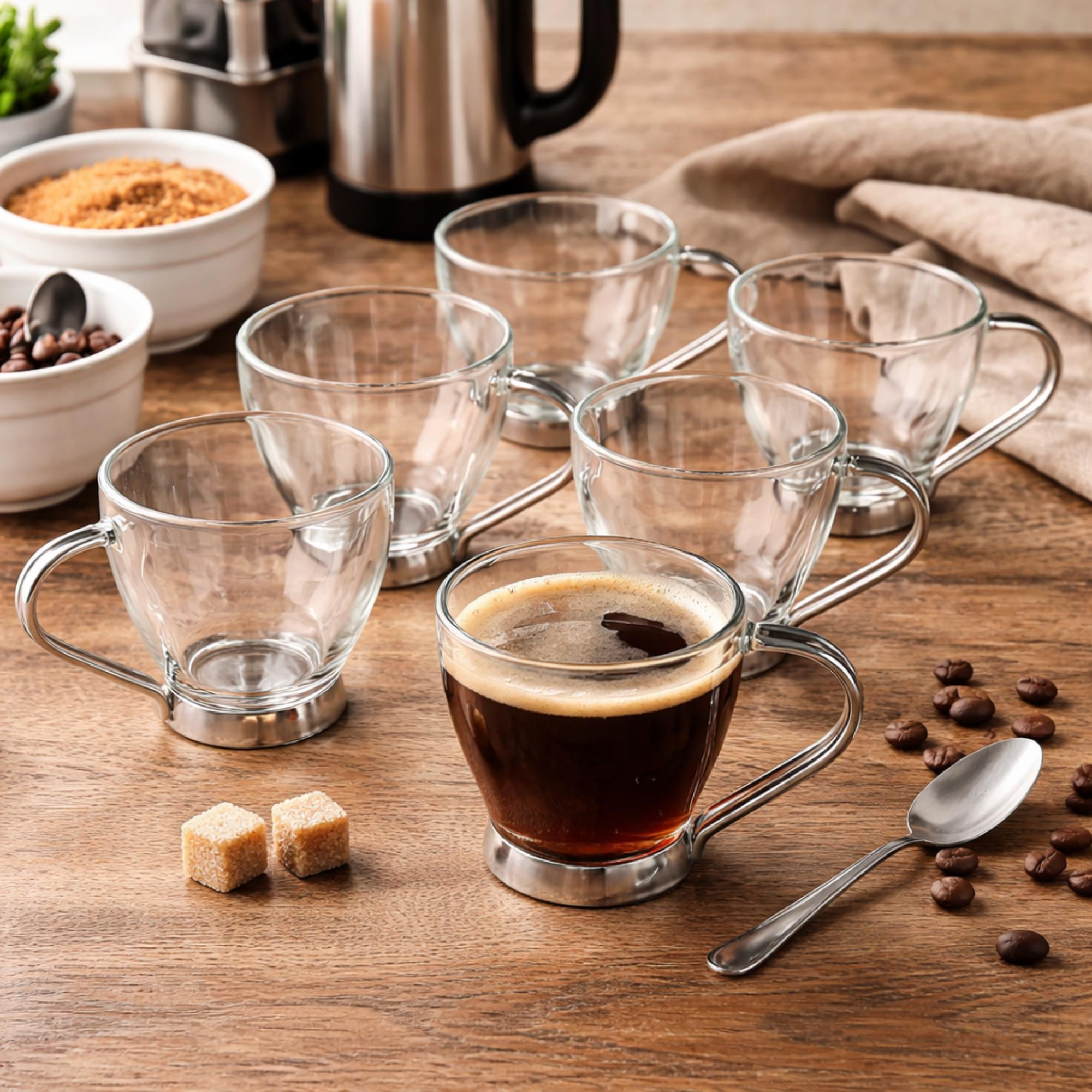 Seis tazas de café de cristal, una llena, sobre mesa de madera con granos y azúcar.