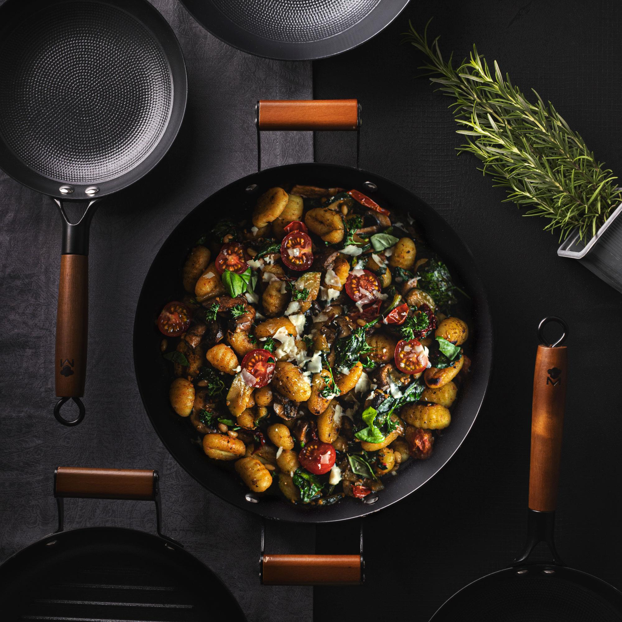 Sartén de gnocchi con tomates cherry, champiñones y hierbas, rodeada de utensilios.