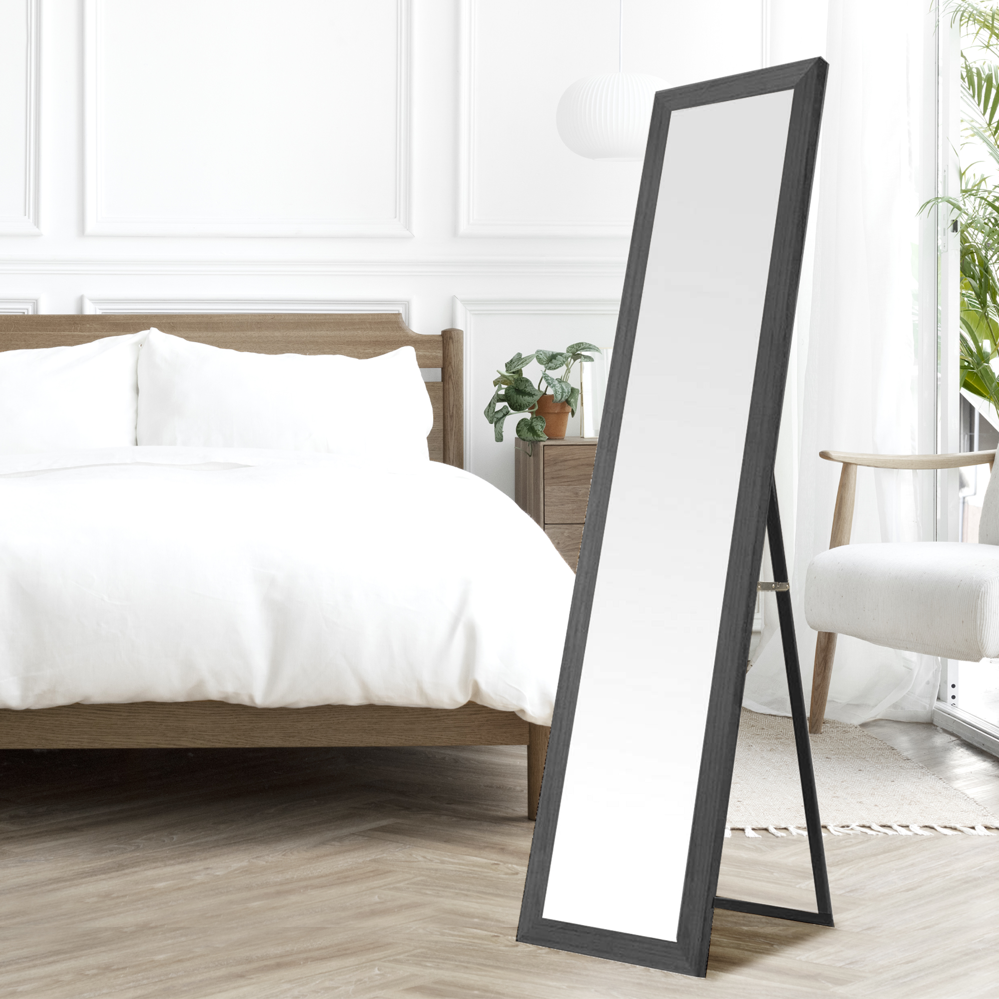 Un espejo alto con marco negro junto a una cama en una habitación luminosa.