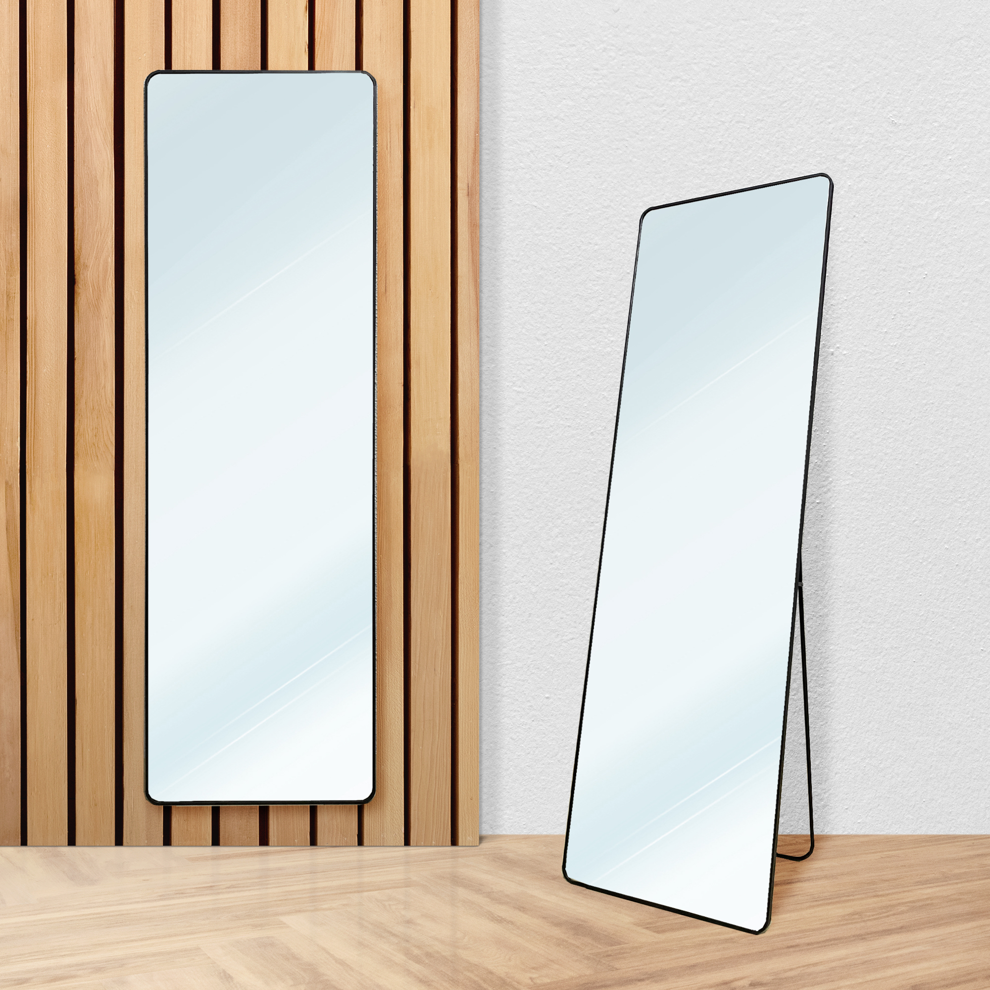 Dos espejos rectangulares con marco negro están sobre suelo de madera frente a pared de madera y pared blanca.