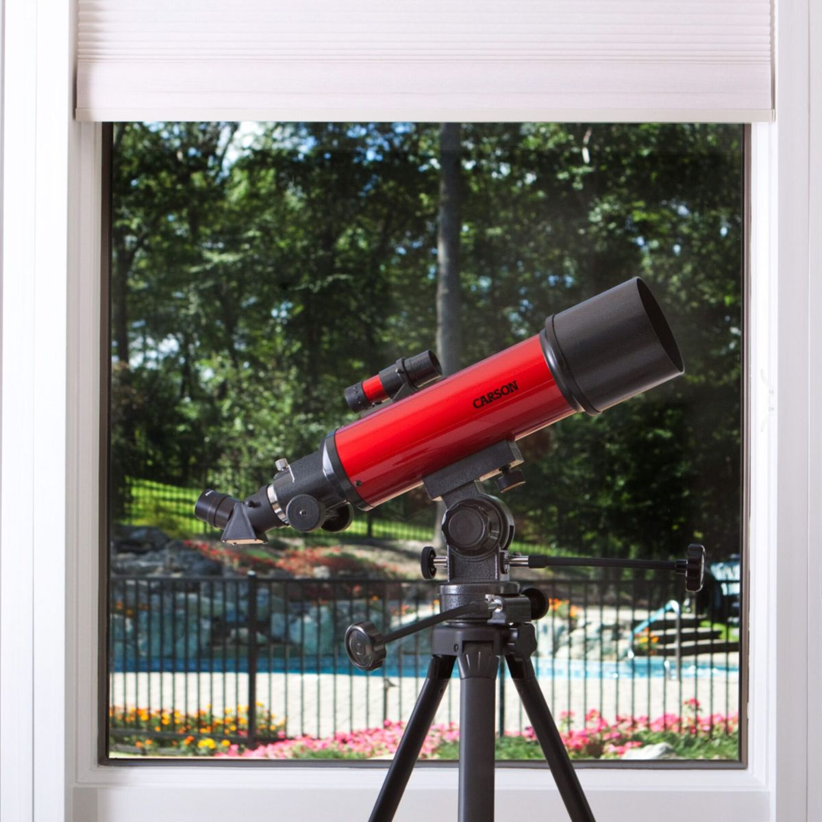 Ein rotes Teleskop auf einem Stativ steht vor einem Fenster mit Blick auf einen Garten.