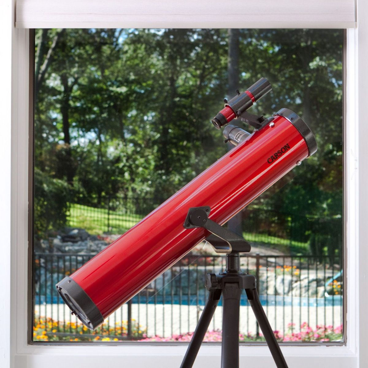 Ein rotes Teleskop steht auf einem Stativ vor einem Fenster mit Blick auf einen Garten und Pool.