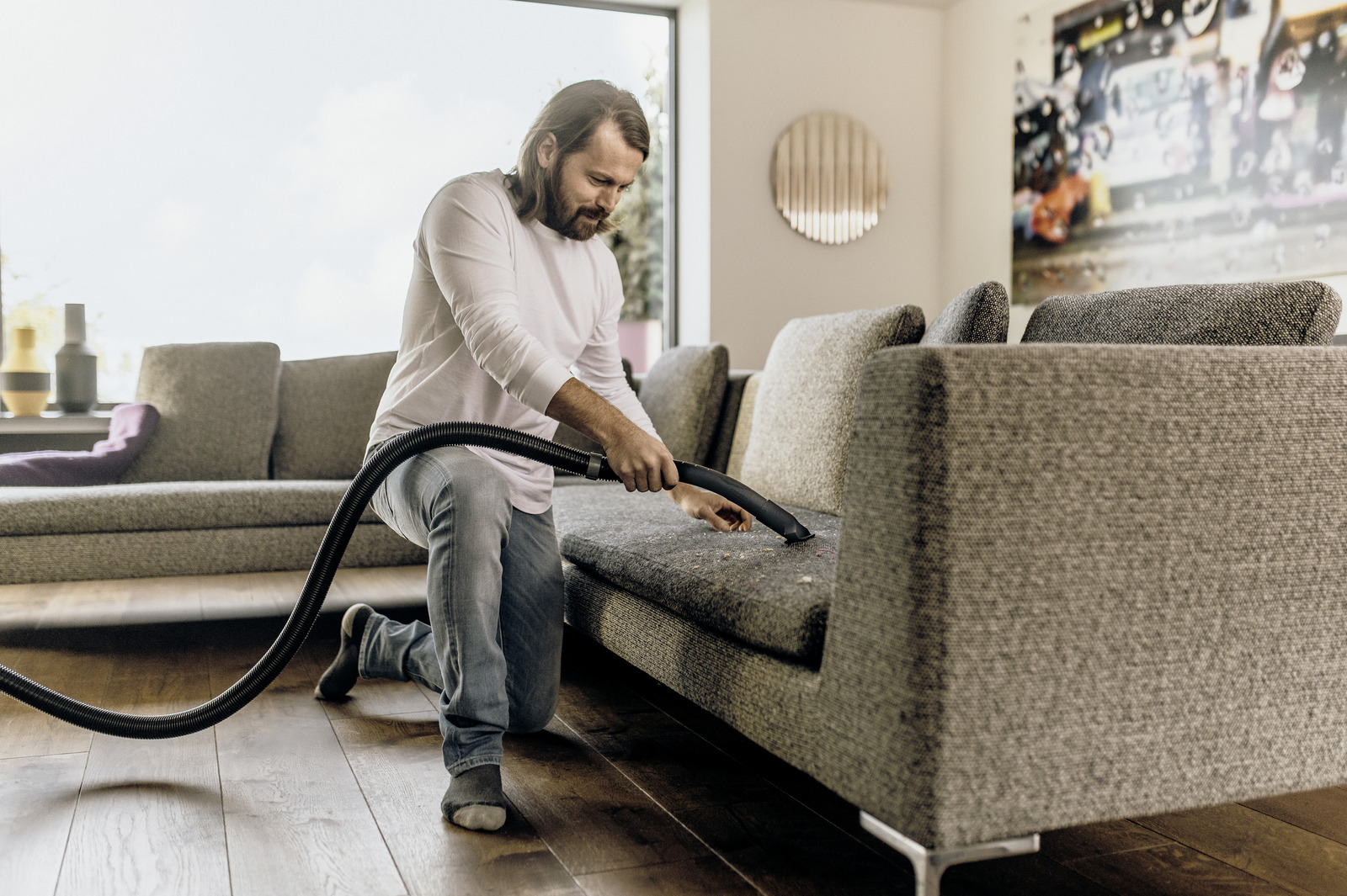 Mężczyzna odkurza sofę, klęcząc na drewnianej podłodze.