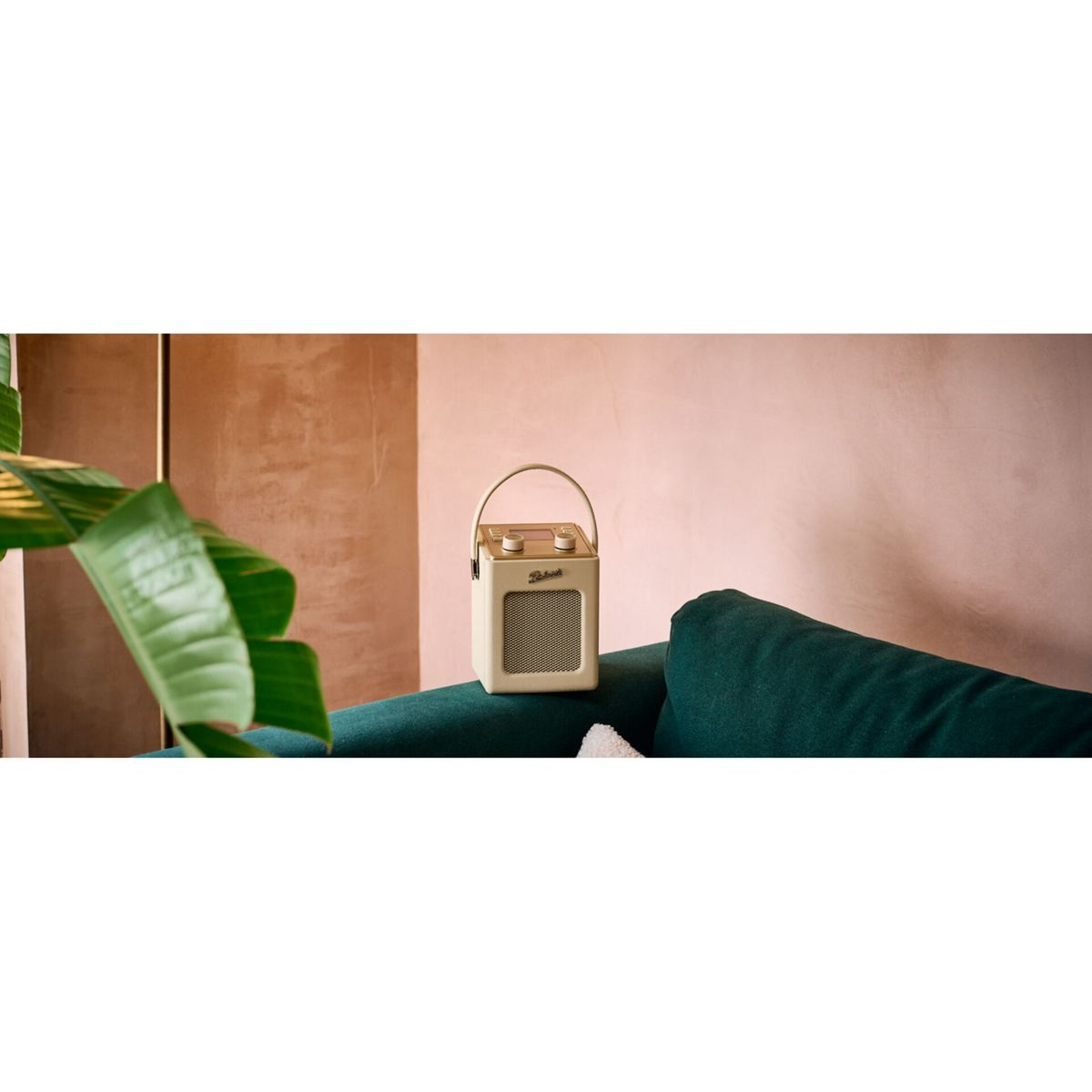Ein cremefarbener Roberts-Radio steht auf einem dunkelgrünen Sofa, vorne ein großes grünes Blatt.