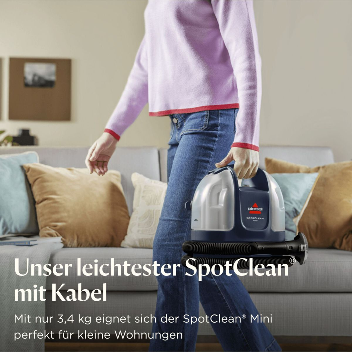Eine Frau trägt einen blauen SpotClean-Reiniger mit Schlauch in einem Wohnzimmer.