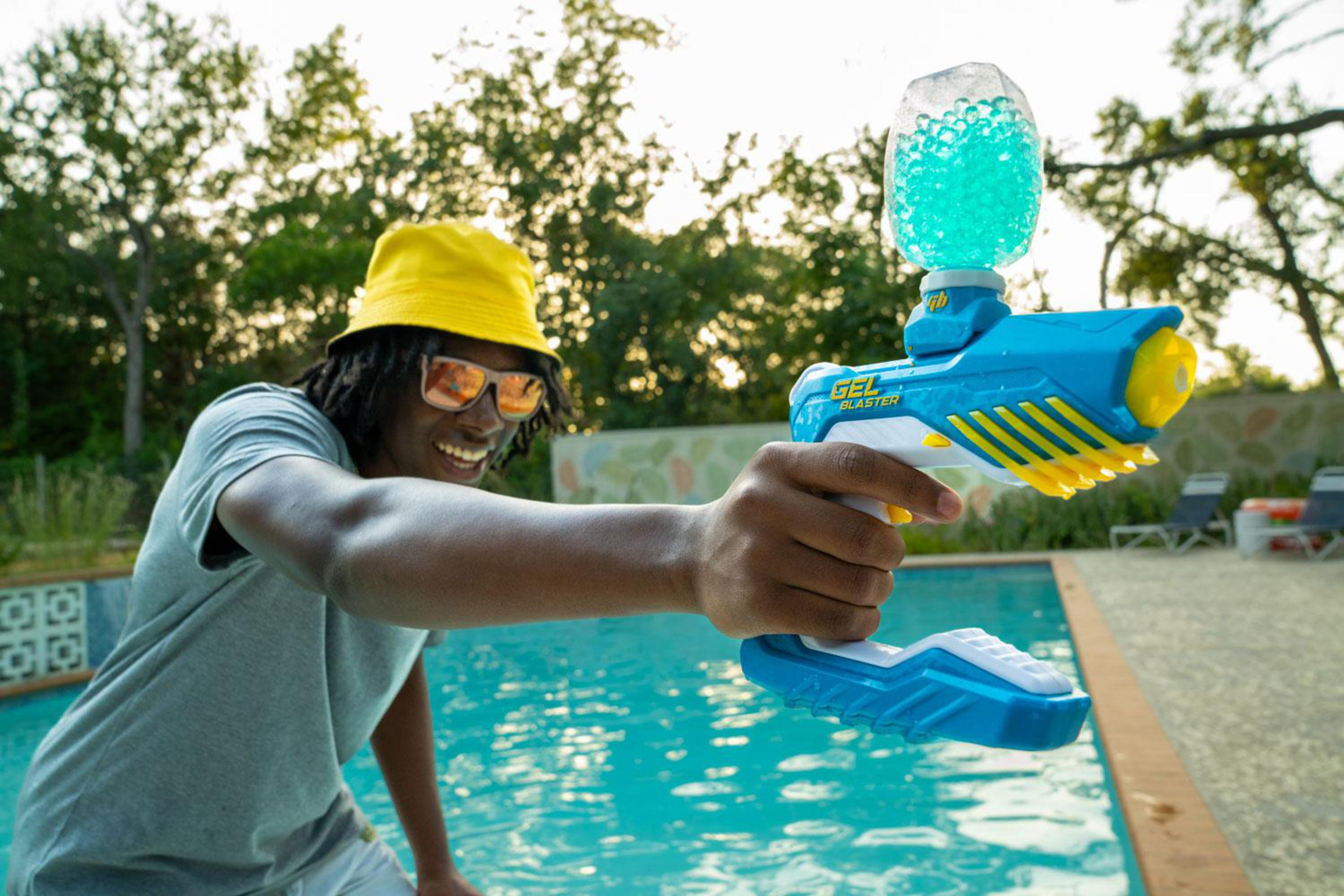Mann mit gelber Mütze und Sonnenbrille hält eine blaue Gel-Blaster-Pistole neben einem Pool.