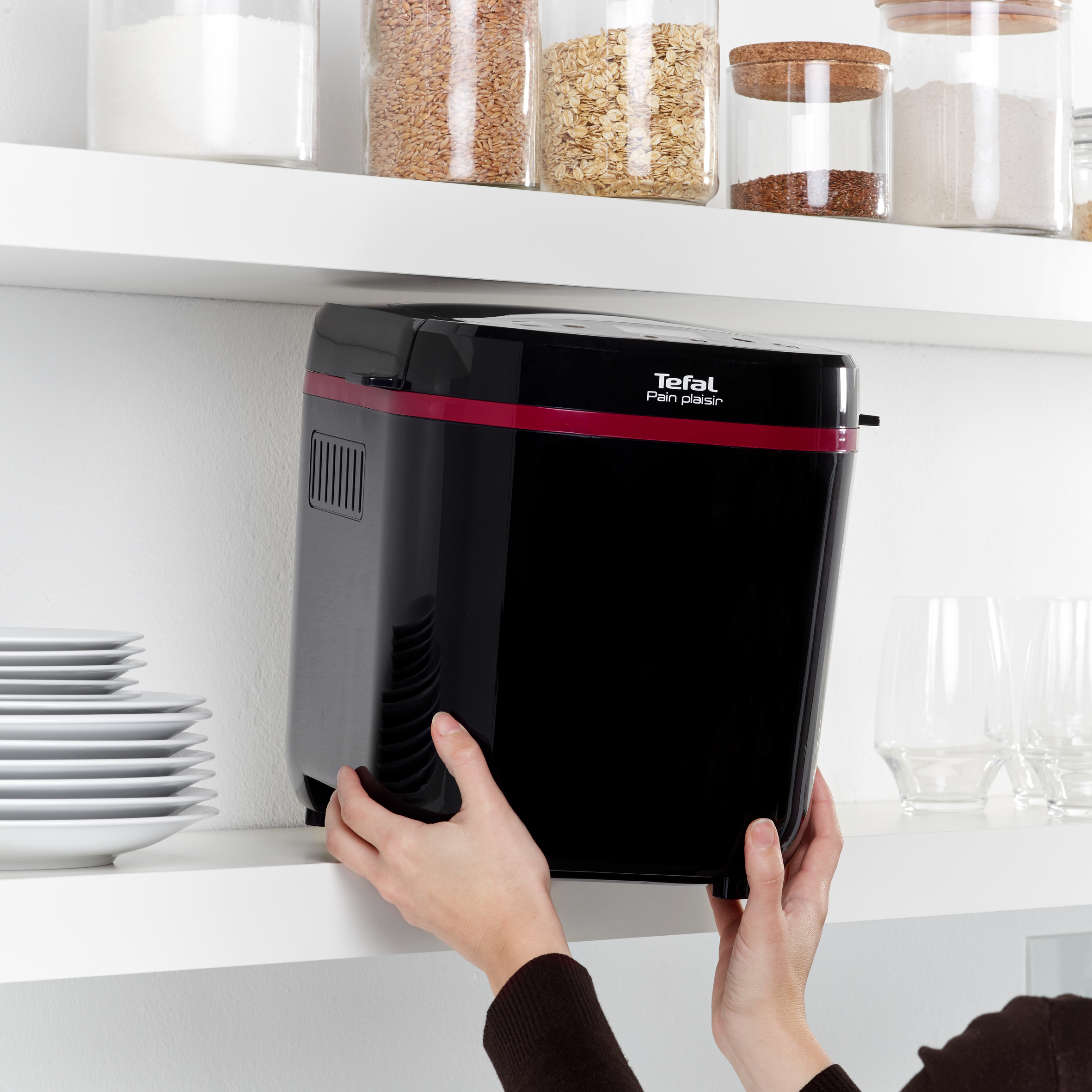 Eine schwarze Brotbackmaschine wird auf ein Regal gestellt, daneben sind Teller und Gläser.