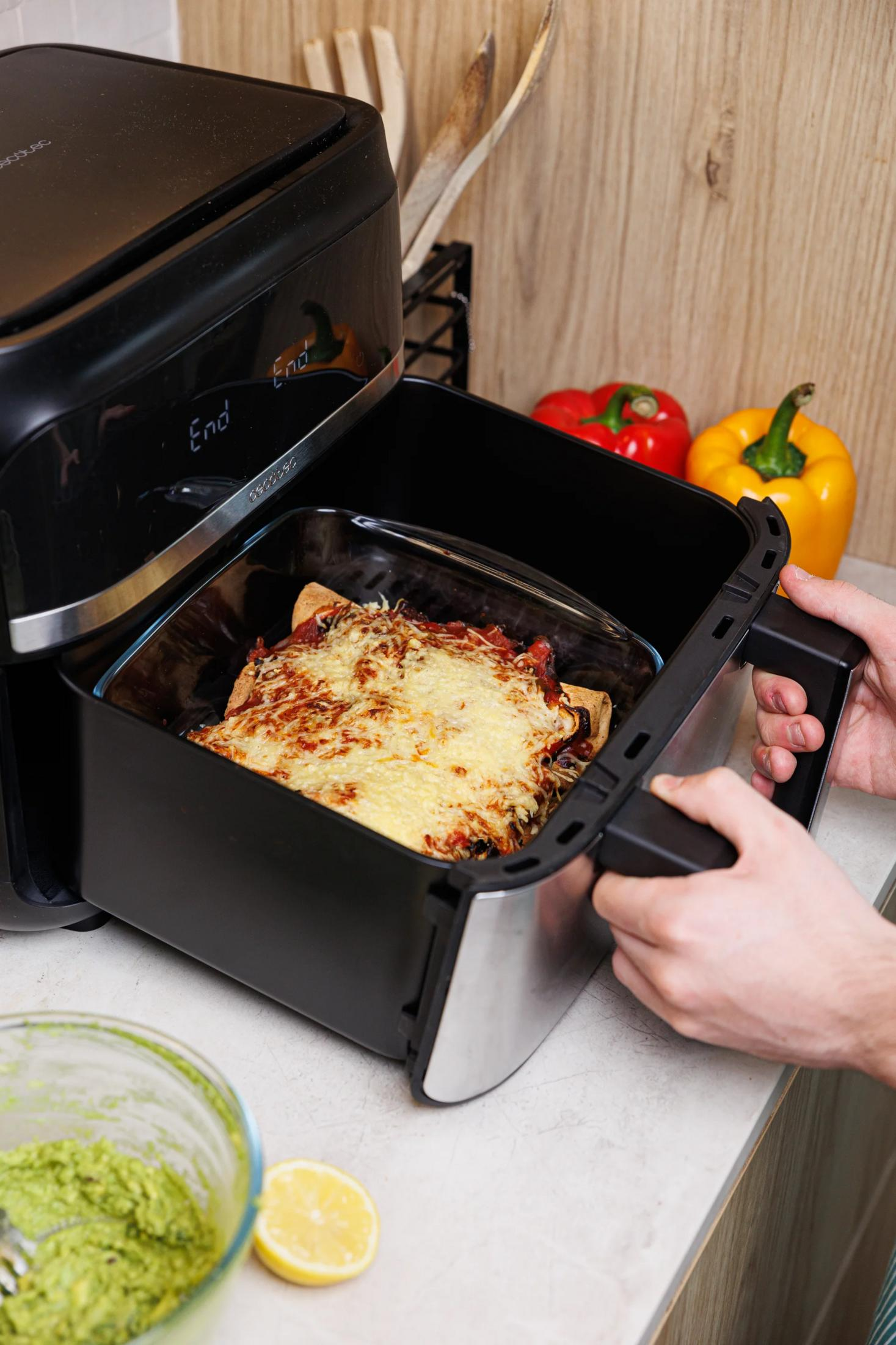 Eine Person nimmt eine Auflaufform mit Käse überbackenem Essen aus einem schwarzen Heißluftfritteuse.