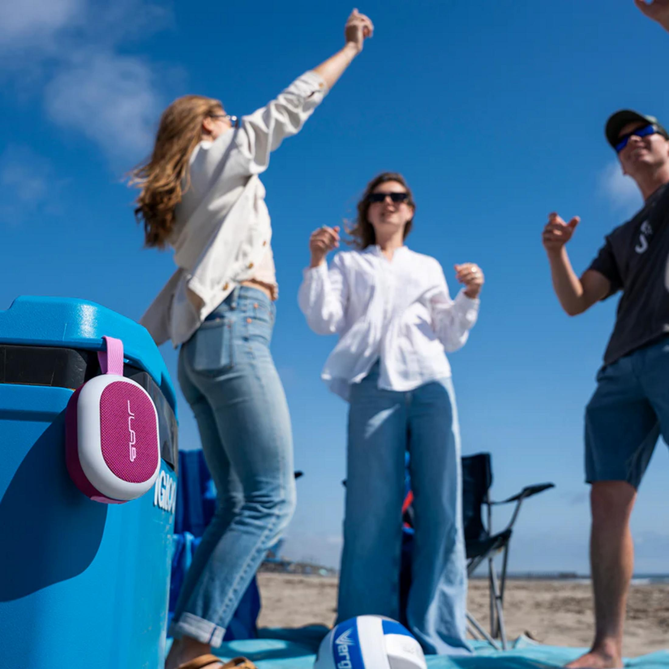 Trois personnes dansent sur la plage. Une petite enceinte rose est accrochée à une poubelle bleue.