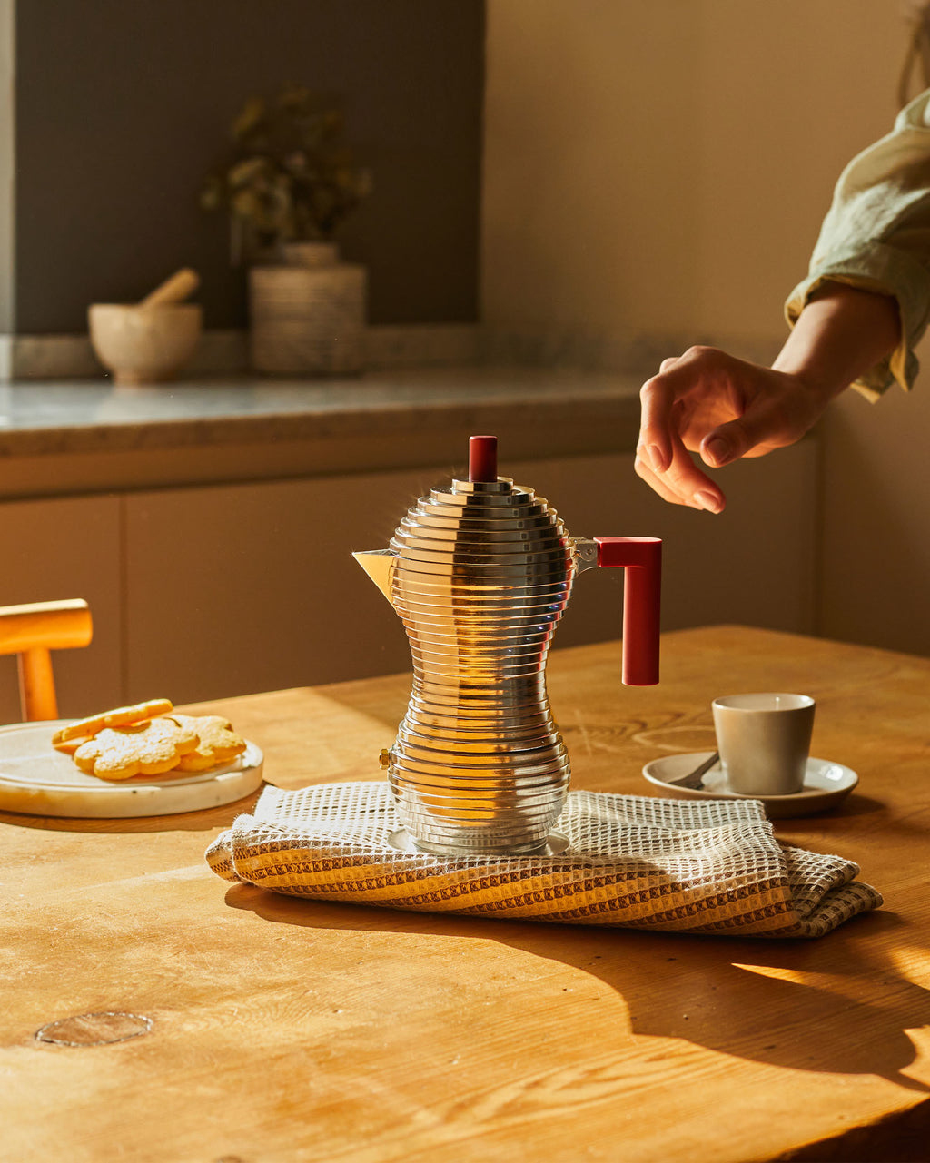 Una caffettiera argentata con manici rossi, biscotti e una tazza di caffè su un tavolo di legno.