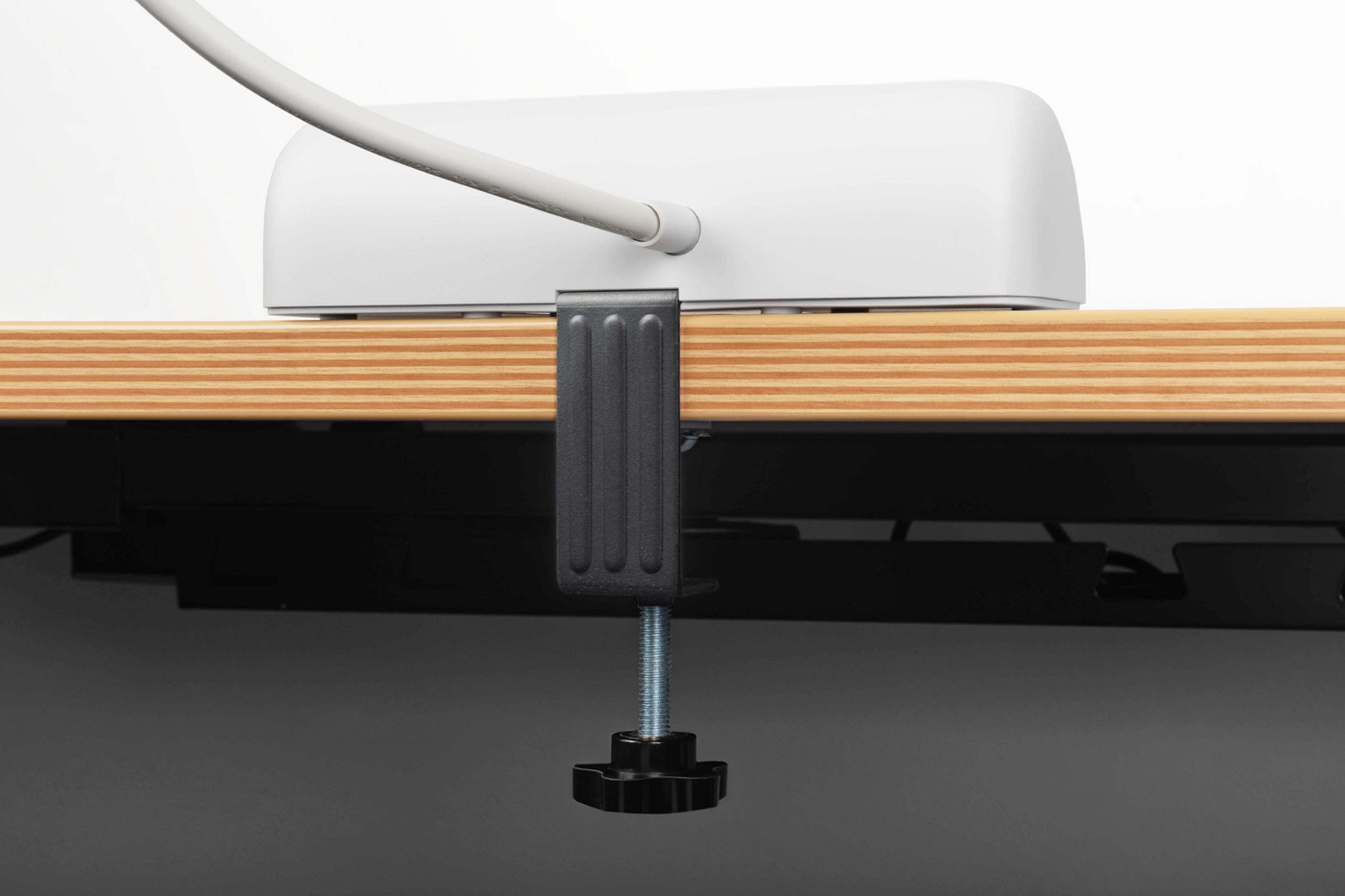Ein weißes Gerät mit Kabel ist an einer Holzplatte befestigt. Dunkler Klemmmechanismus.