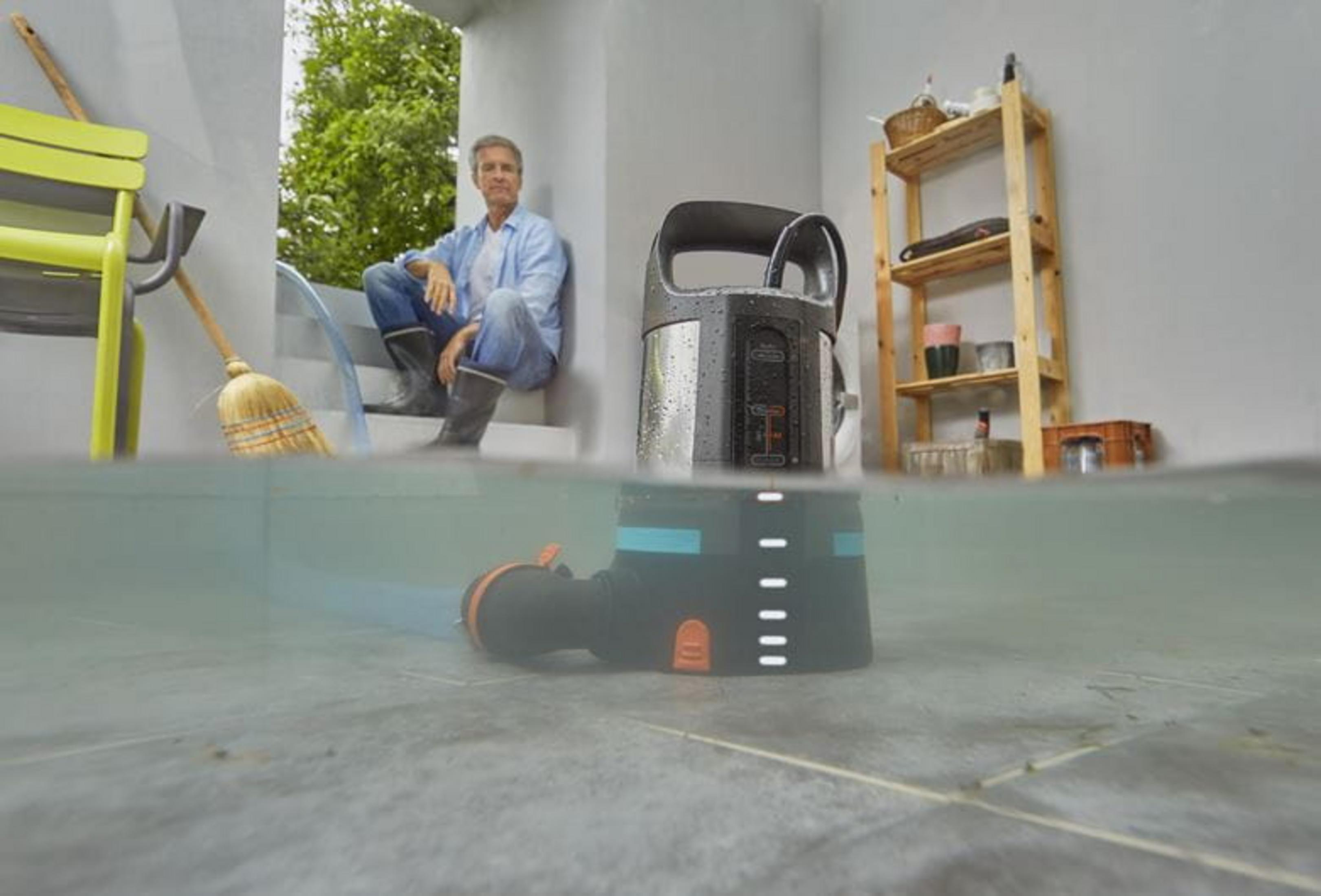 Mann sitzt neben Pumpe in Wasser. Besen, Stuhl, Regal im Hintergrund.