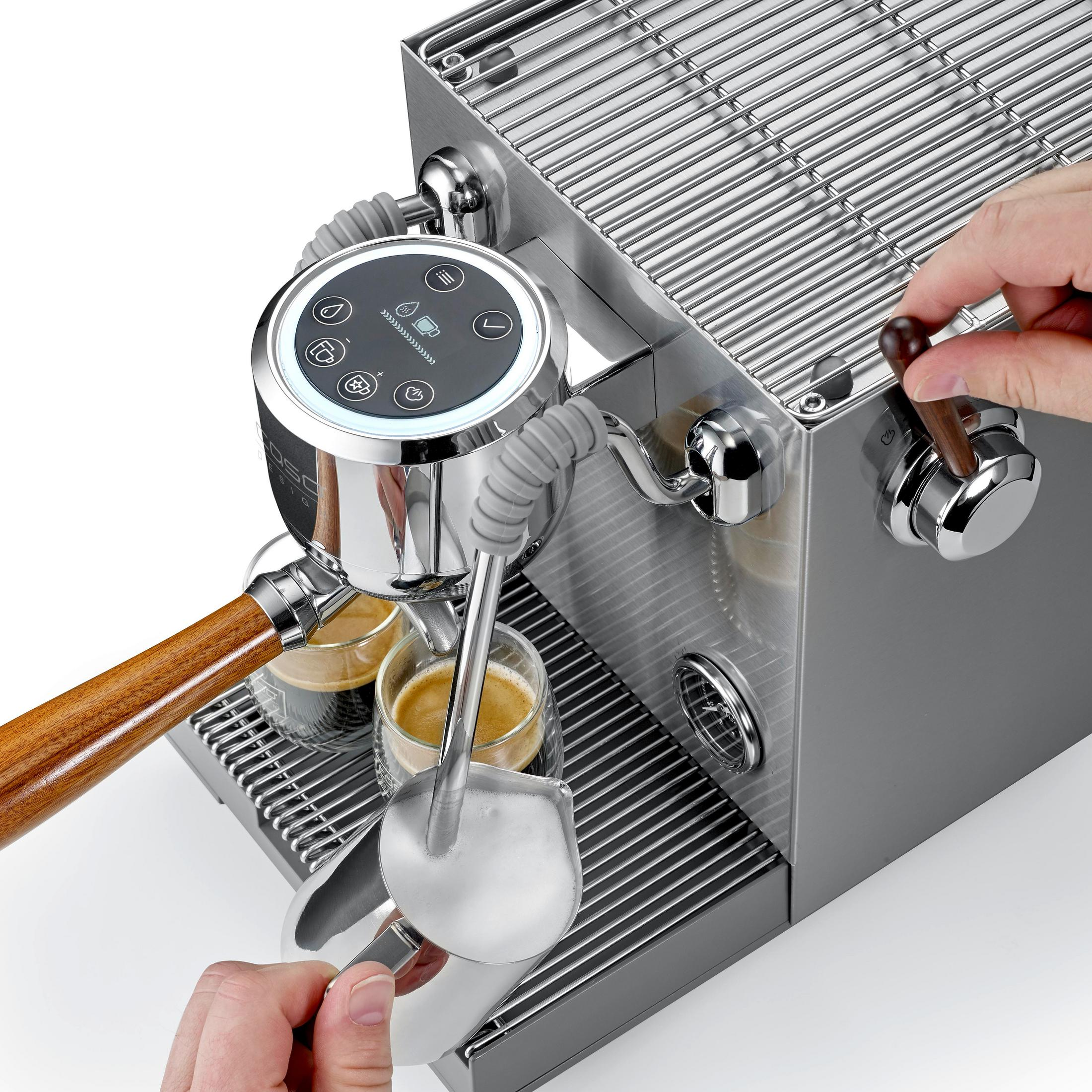 Eine Espressomaschine mit Holzgriff brüht Kaffee in zwei Gläser. Eine Hand schäumt Milch auf.