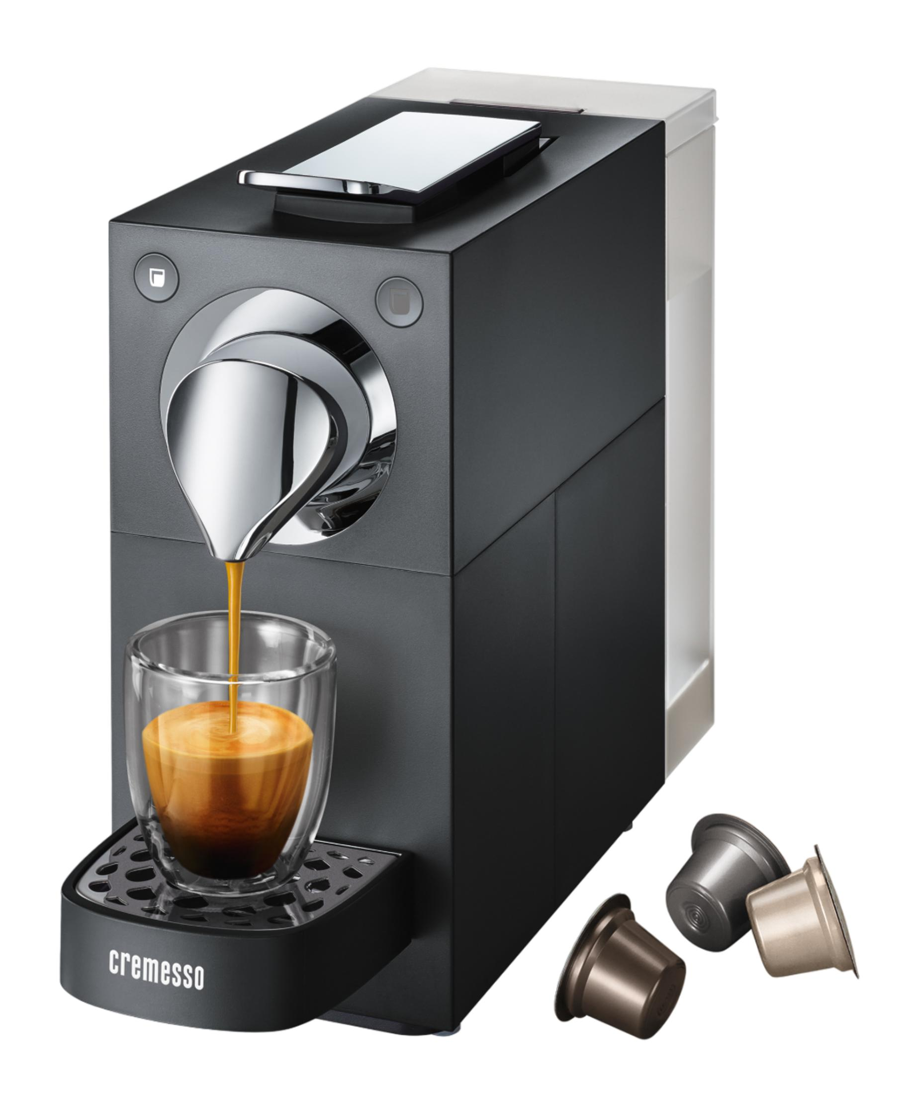 Schwarze Kaffeemaschine brüht Espresso in ein Glas; drei Kapseln liegen daneben.