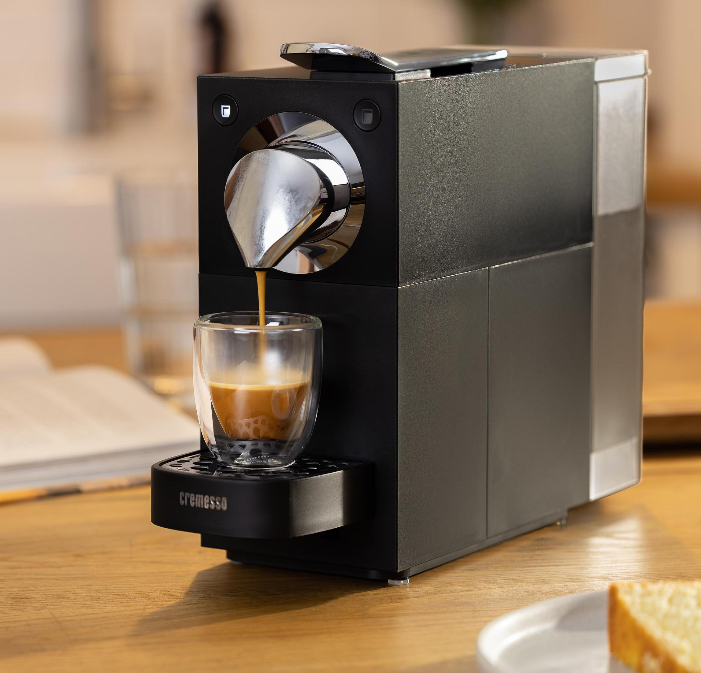 Eine schwarze Kaffeemaschine brüht Espresso in ein doppelwandiges Glas auf einem Holztisch.