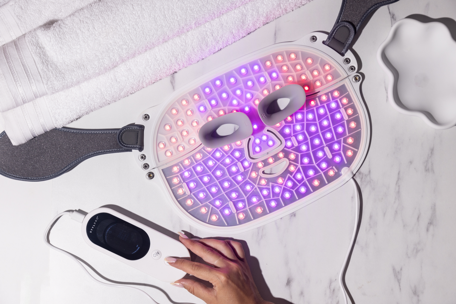 Un dispositivo per maschera facciale a LED con luci rosse e viola, accanto a un asciugamano arrotolato bianco.