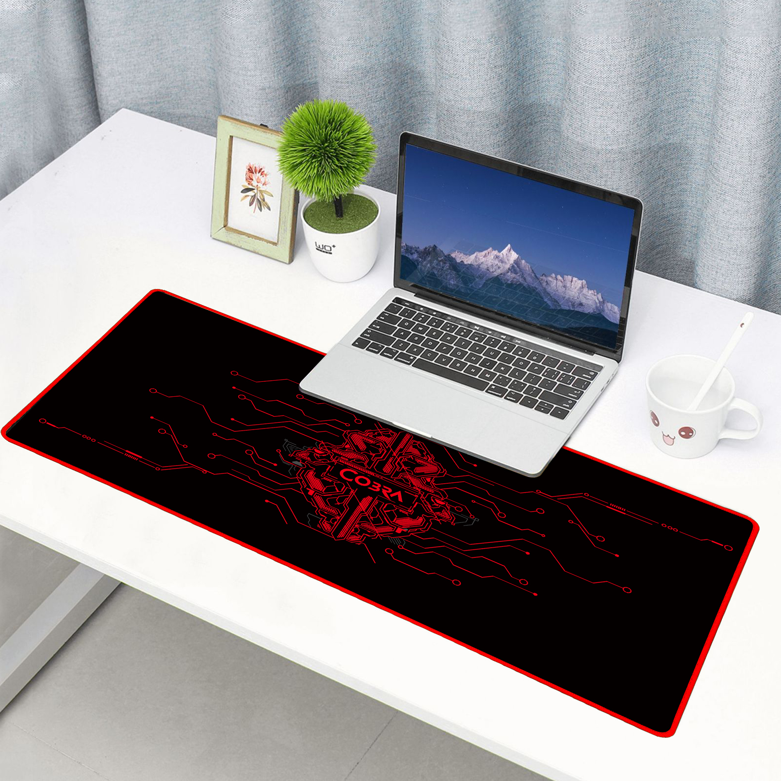 Laptop na czarnej podkładce pod mysz z czerwonym wzorem obwodu. Obok ramka ze zdjęciem i roślina.