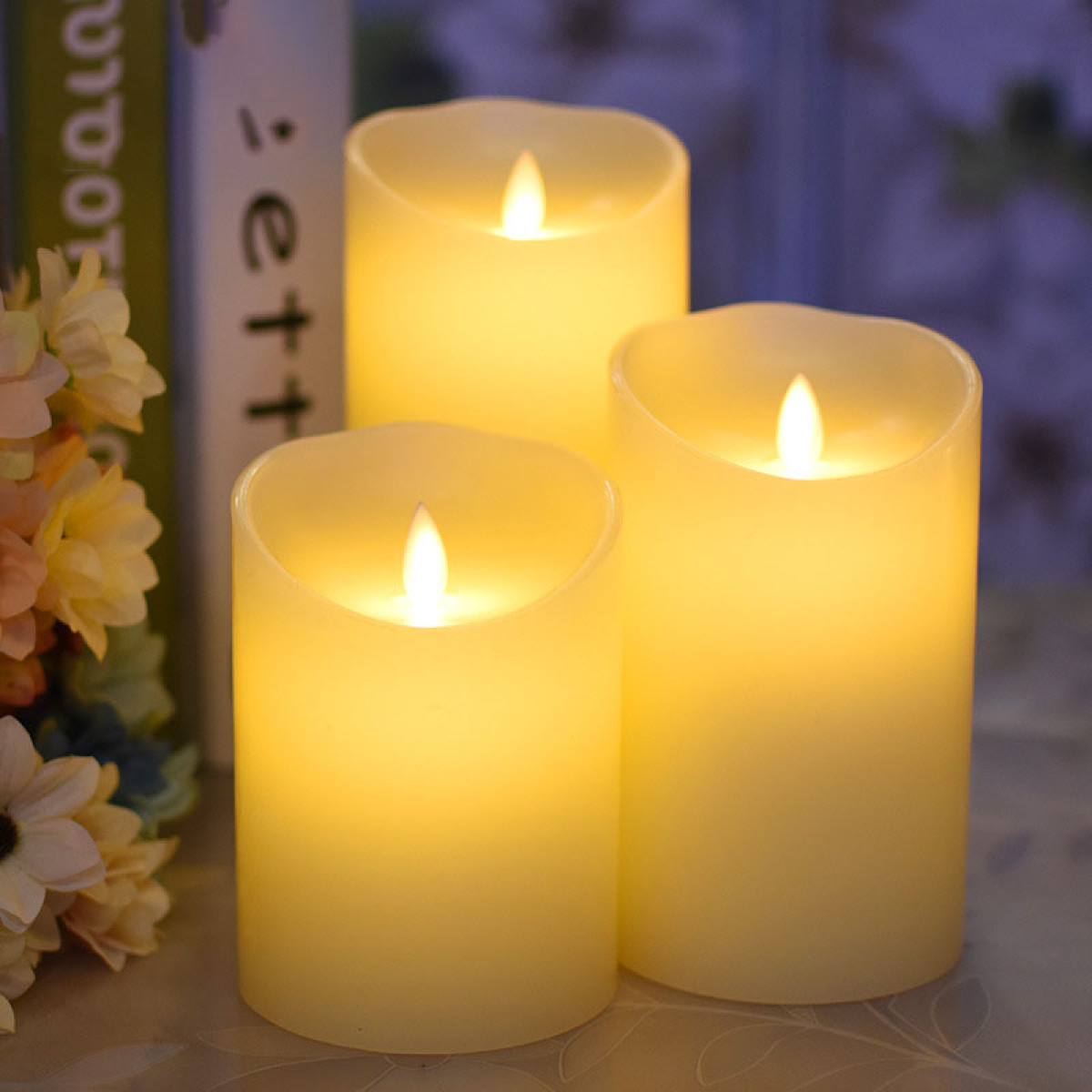Trois bougies LED crème avec flammes brillent, près de livres et fleurs.