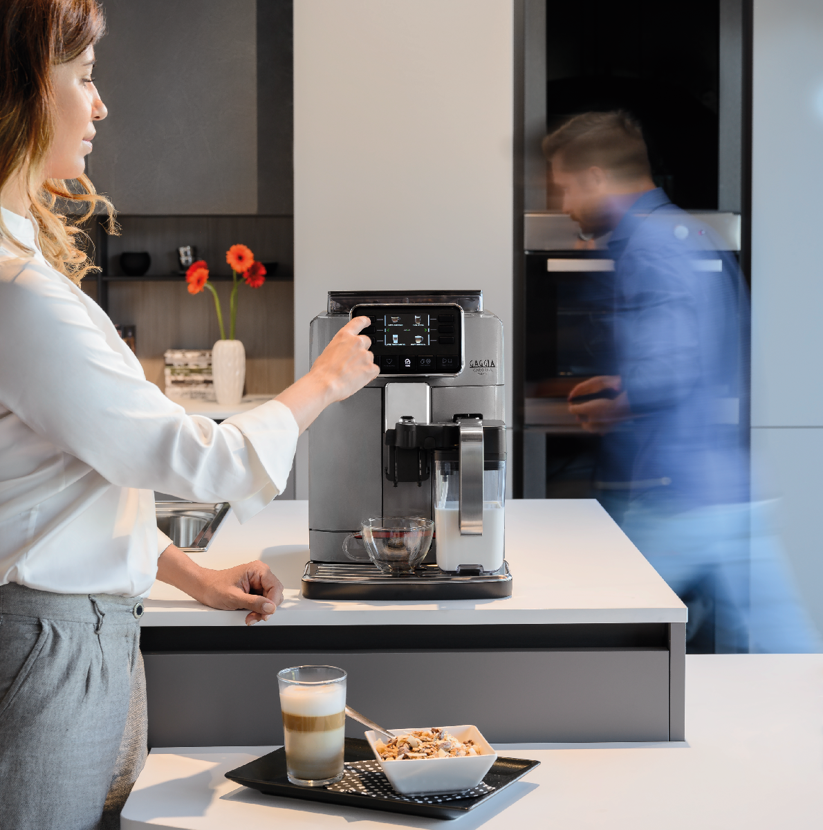 Una donna usa una macchina da caffè in cucina. Un uomo è sfocato sullo sfondo.