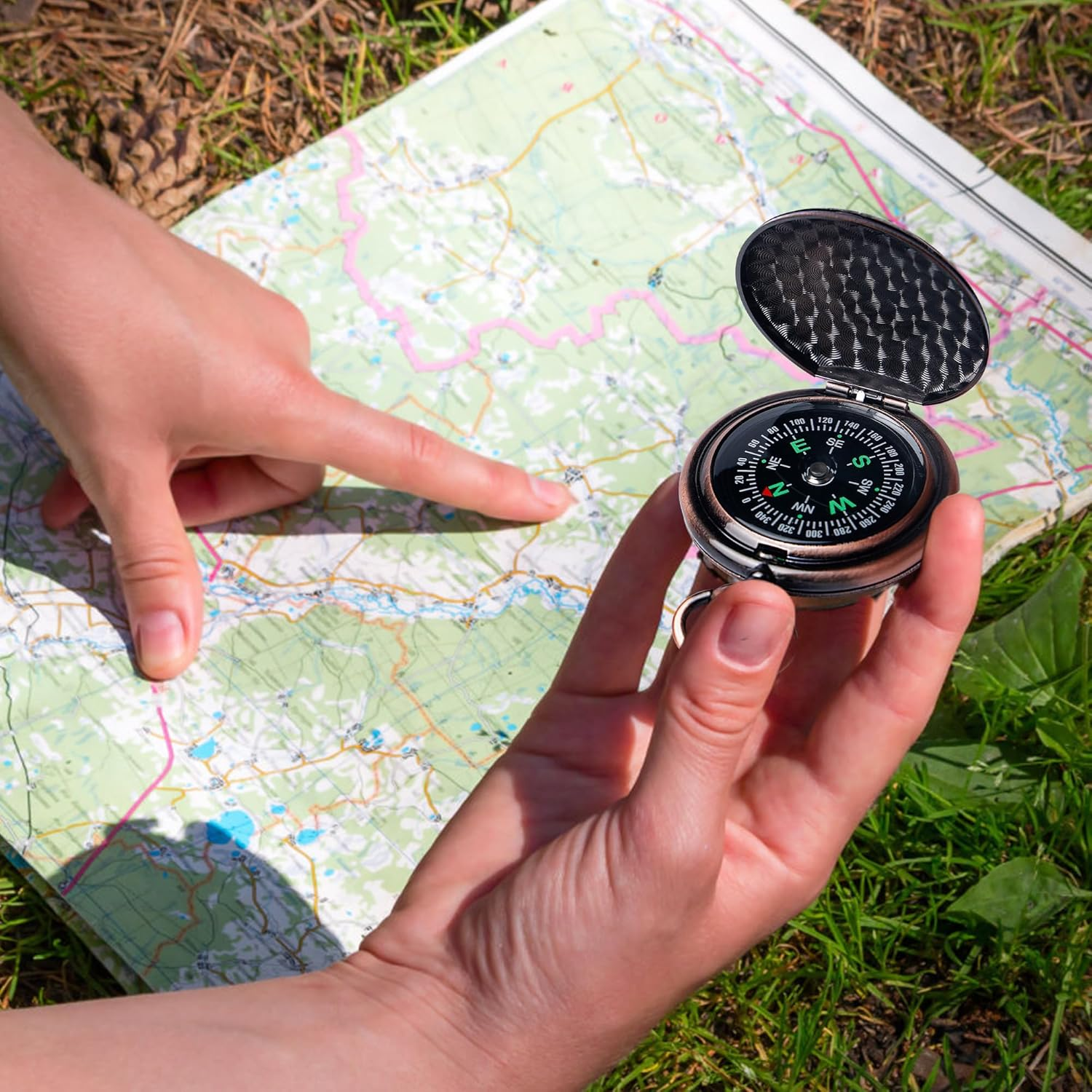 Hände halten einen Kompass über eine Karte auf Gras. Ein Finger zeigt auf die Karte.