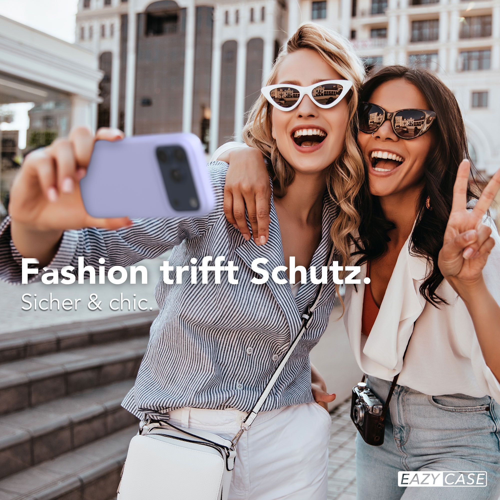 Zwei lachende Frauen machen ein Selfie mit einem lilafarbenen Handy vor einem Gebäude.