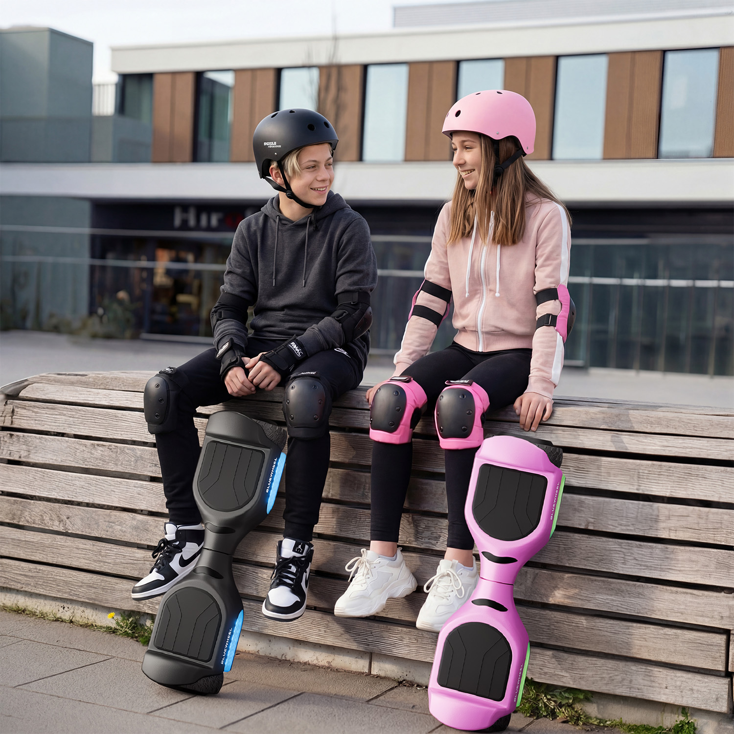 Zwei Kinder mit Schutzausrüstung sitzen auf einer Bank neben Hoverboards.