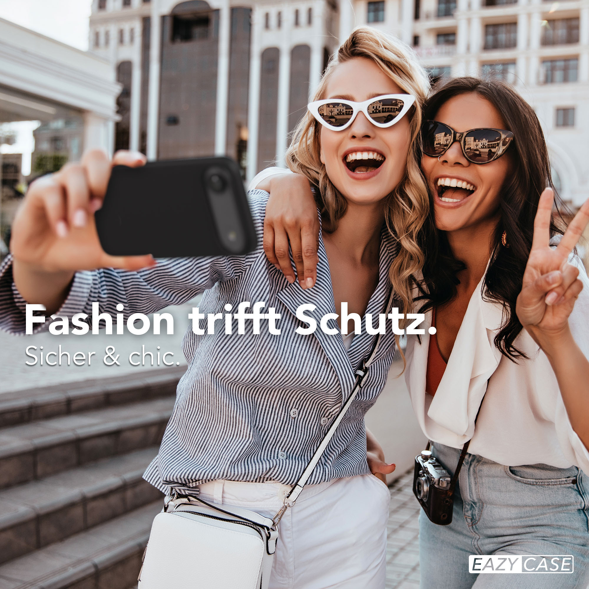 Zwei lachende Frauen machen ein Selfie vor einem Gebäude, eine hält ein Handy.
