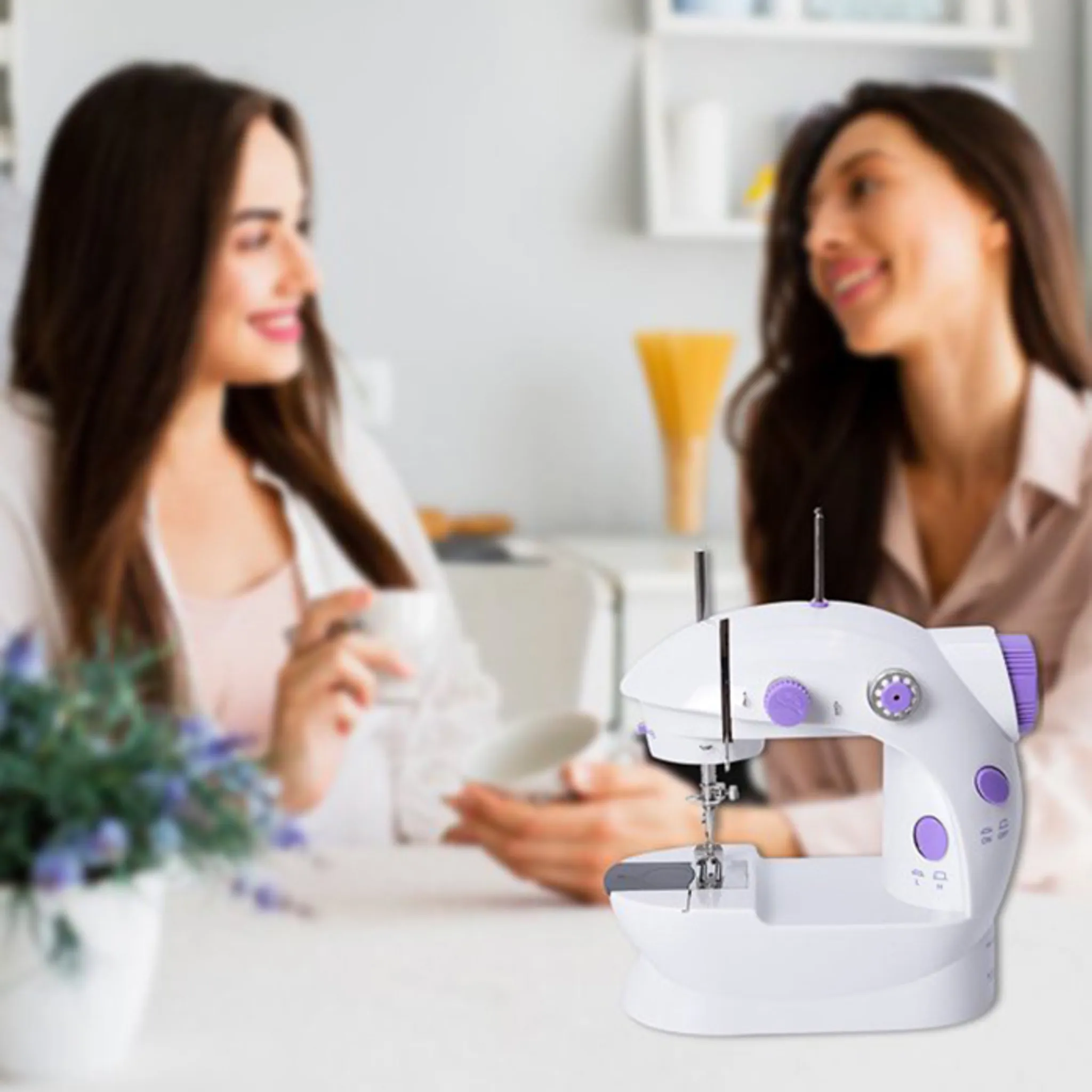 Eine weiße Nähmaschine mit lila Akzenten steht im Vordergrund. Zwei Frauen sind unscharf im Hintergrund zu sehen.