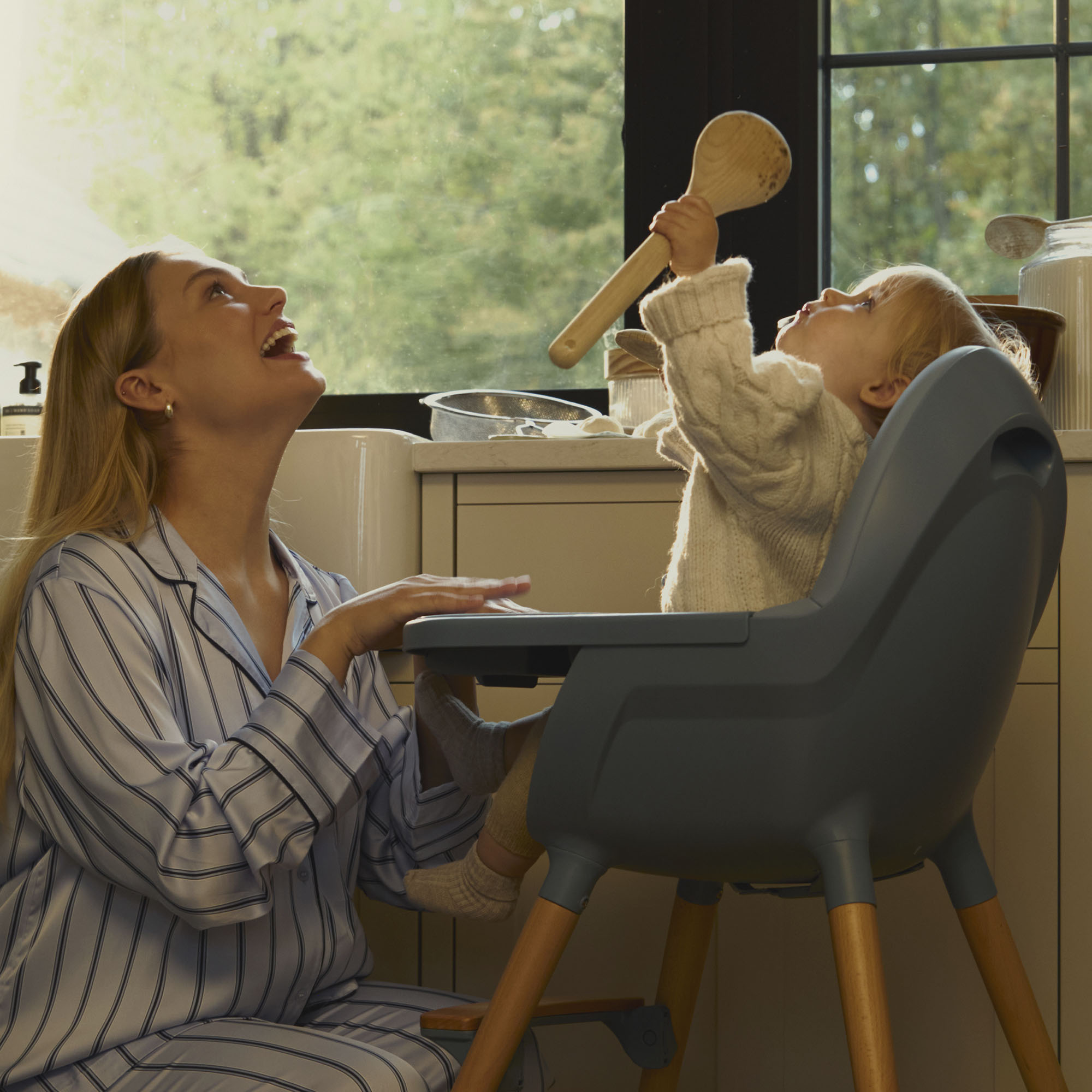 Mutter lacht, während Baby Löffel hält. Küche im Hintergrund.