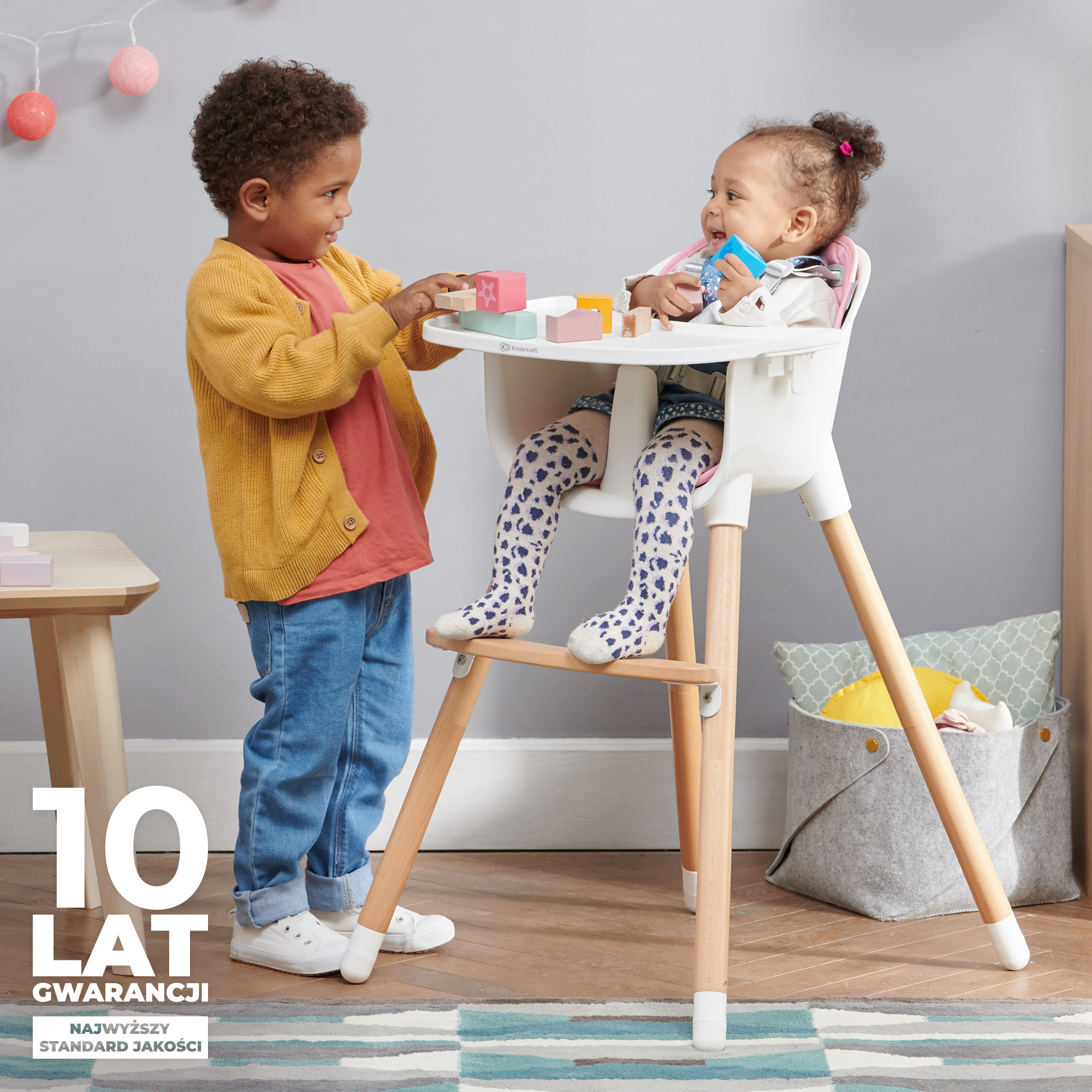 Zwei Kinder spielen mit Bauklötzen. Ein Kind sitzt in einem Hochstuhl, das andere steht daneben.