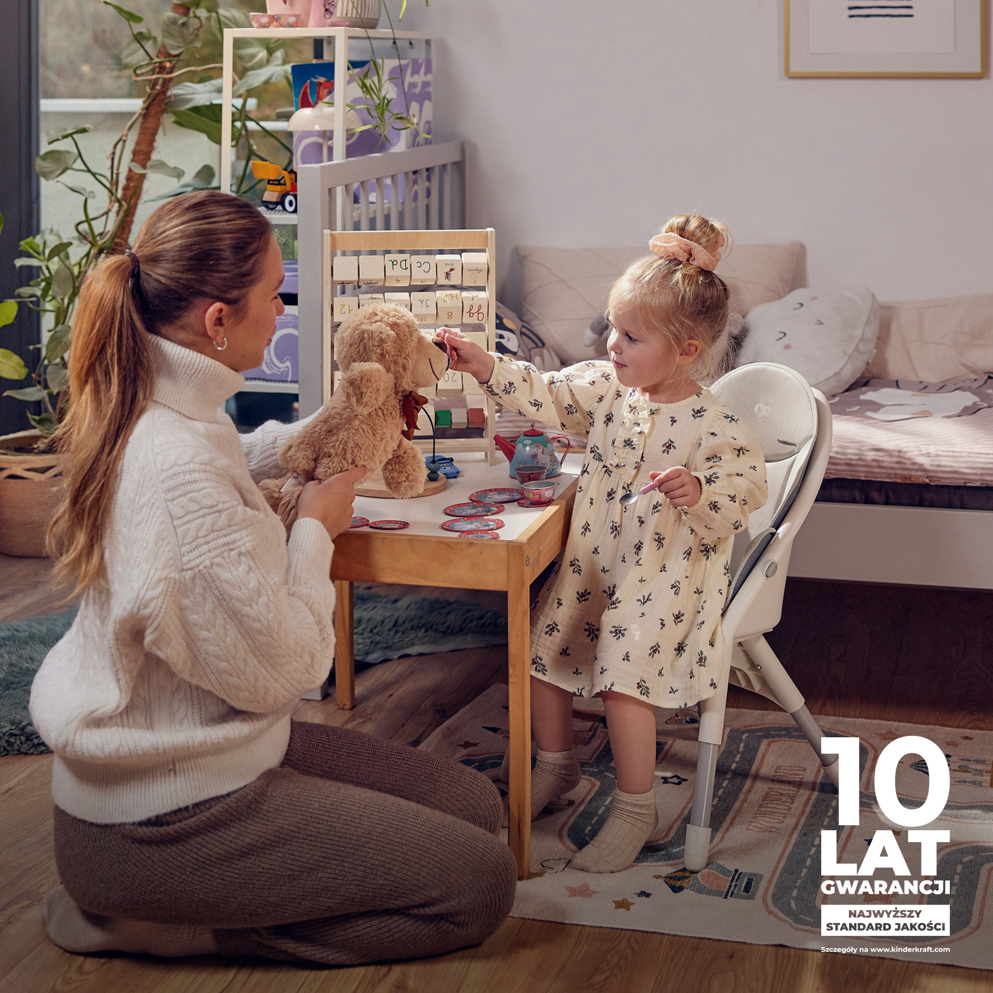 Eine Frau kniet und spielt mit einem Teddybären mit einem kleinen Mädchen an einem Holztisch.