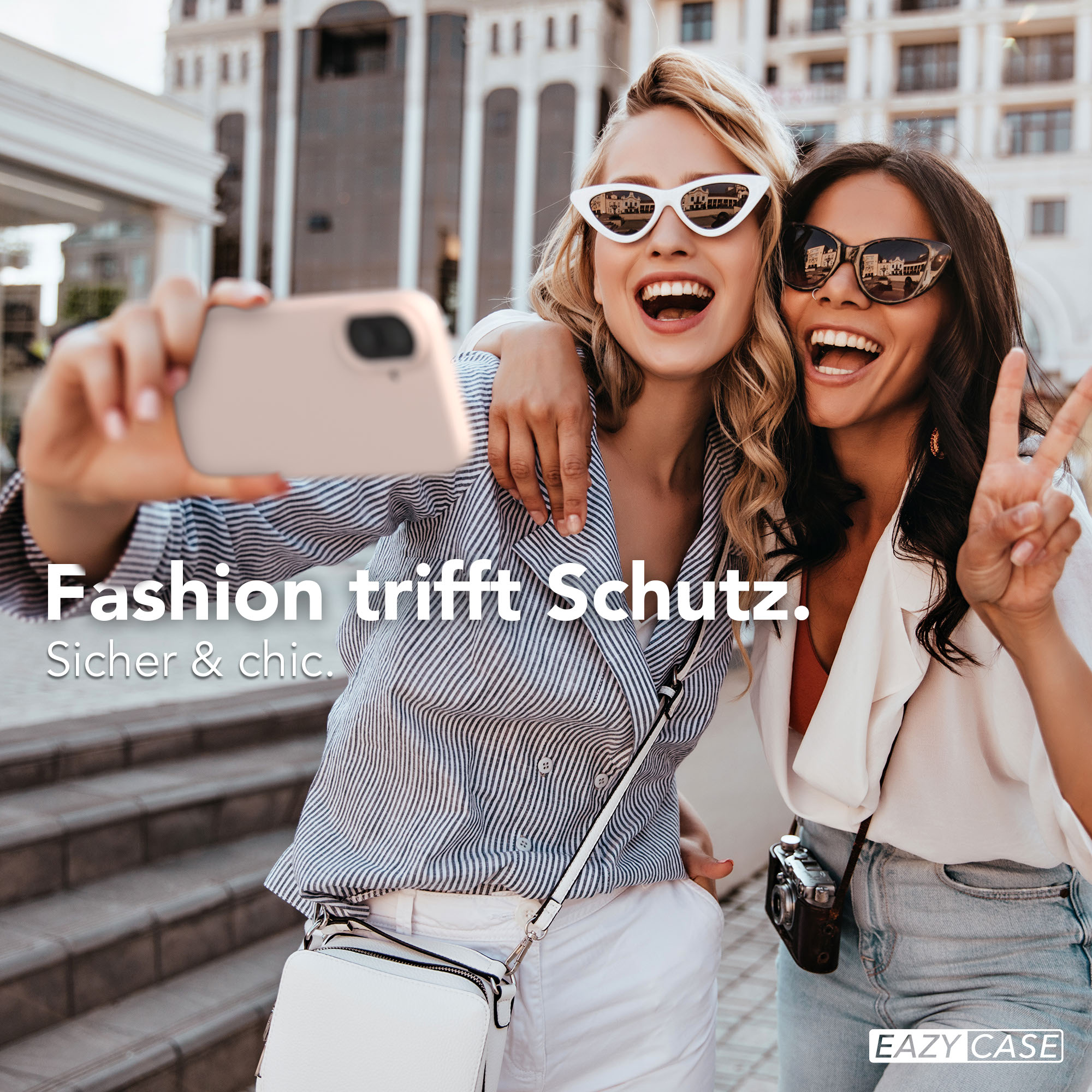 Zwei lachende Frauen machen ein Selfie vor einem Gebäude. Eine winkt mit der Hand.