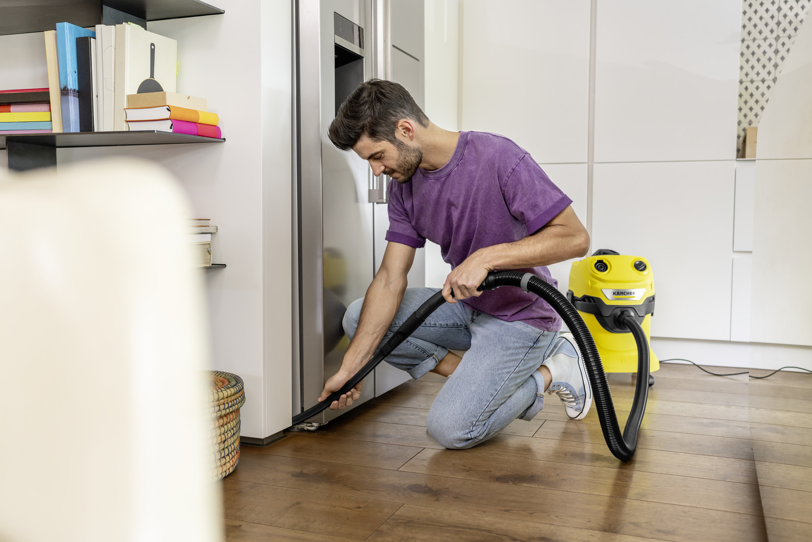 Hombre con camisa morada aspira suelo cerca de nevera con aspiradora Karcher amarilla.