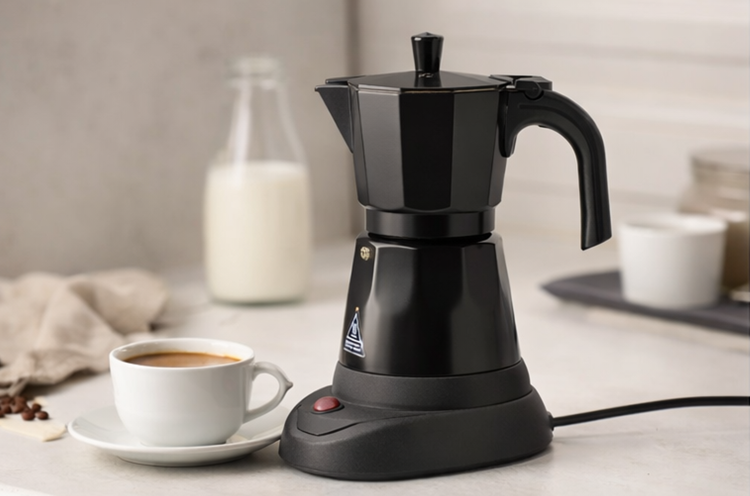 Una cafetera moka negra junto a una taza de café, con una botella de leche al fondo.