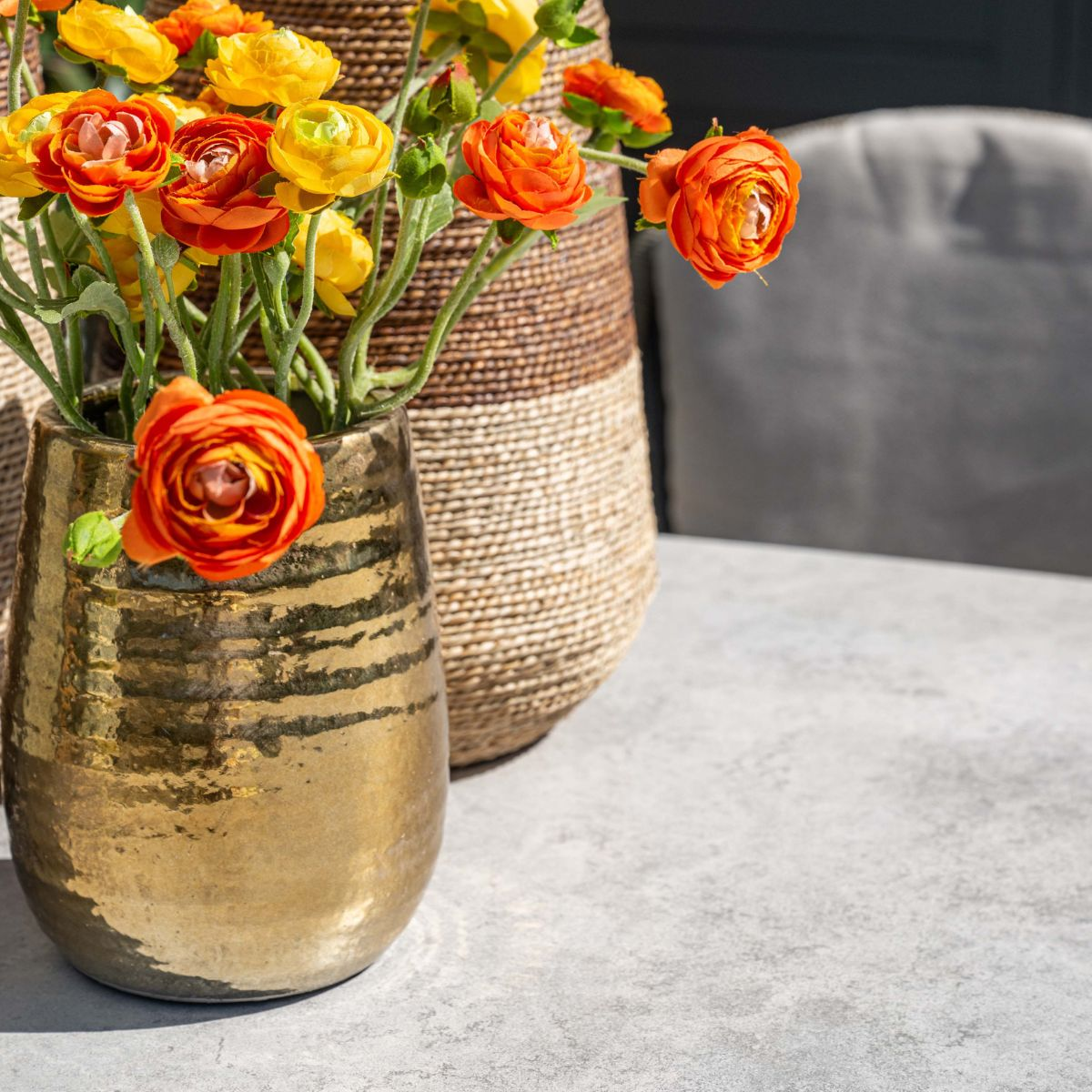 Oranje en gele ranonkels in een metallic gouden vaas, met erachter een gevlochten mand.