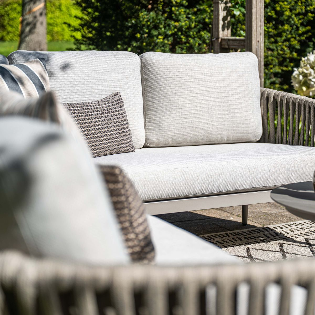 A beige outdoor sofa with cushions on a stone patio, with green bushes in the background.
