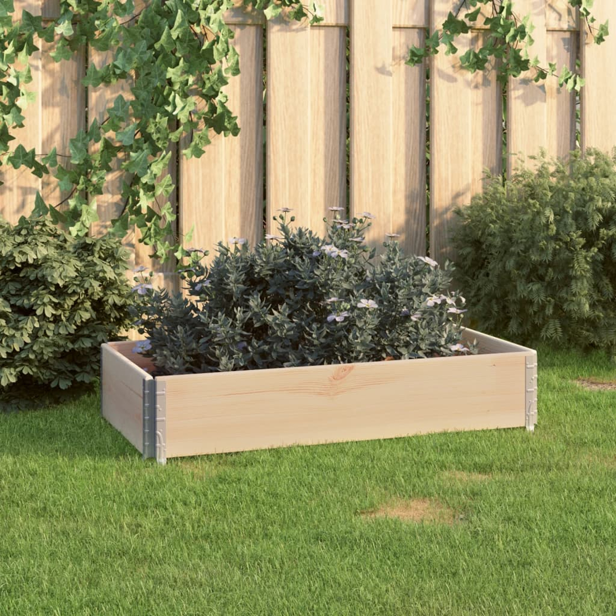 Ein heller Holzpflanzkasten mit grünen Pflanzen und kleinen weißen Blüten steht auf grünem Gras vor einem Holzzaun.