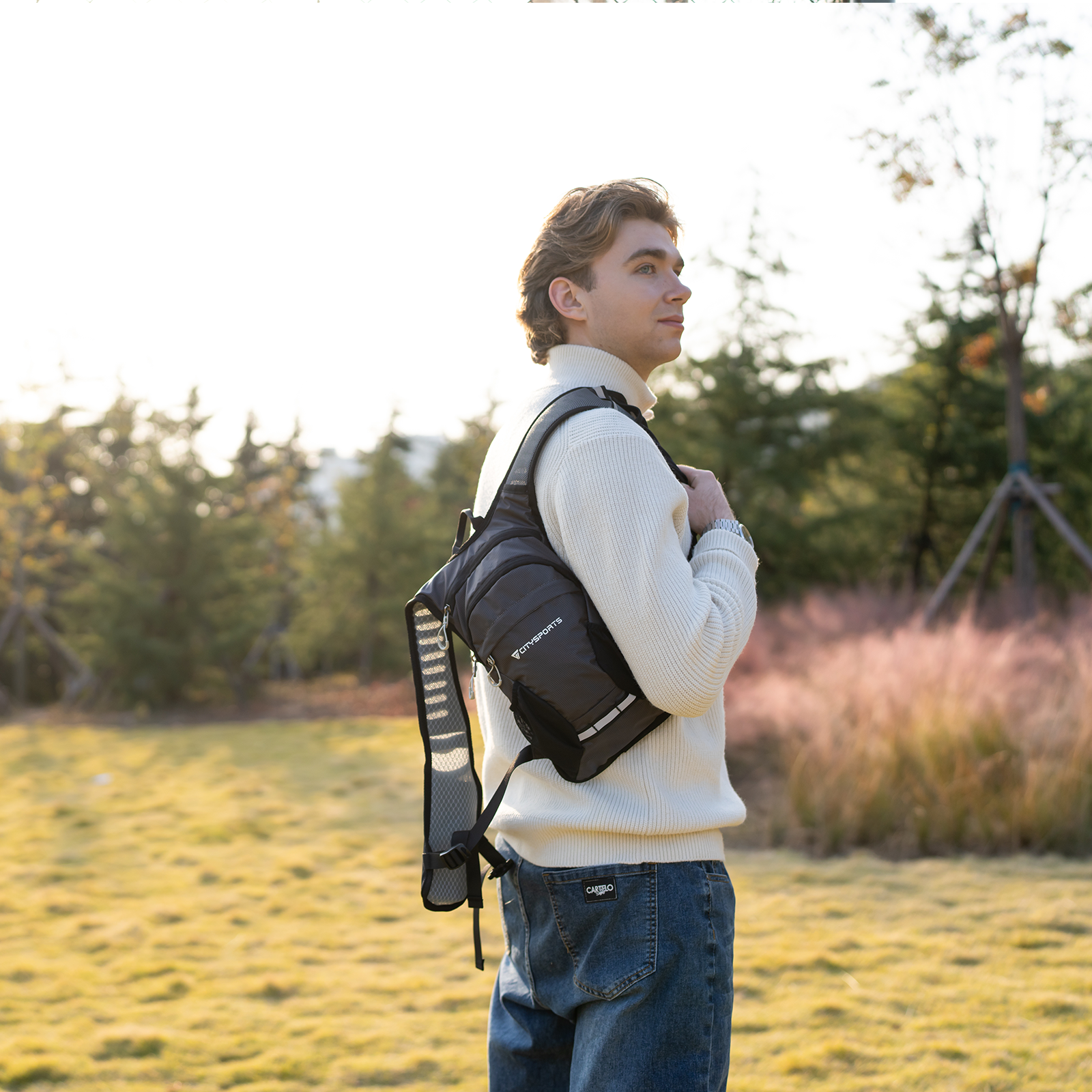 Mężczyzna w białym swetrze i dżinsach nosi szary plecak na zewnątrz.