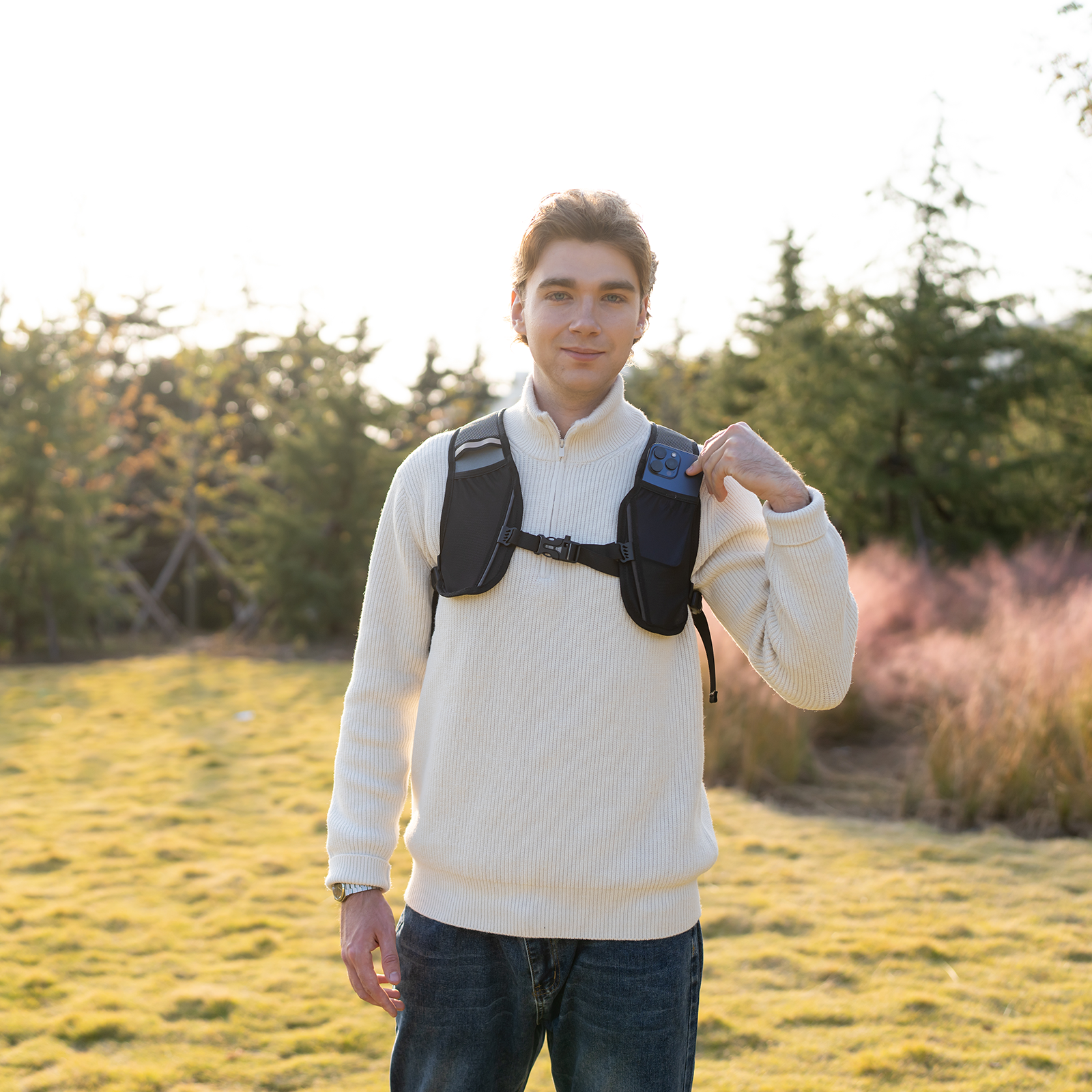 Mężczyzna w białym swetrze z kamizelką i telefonem na zewnątrz o zachodzie słońca.