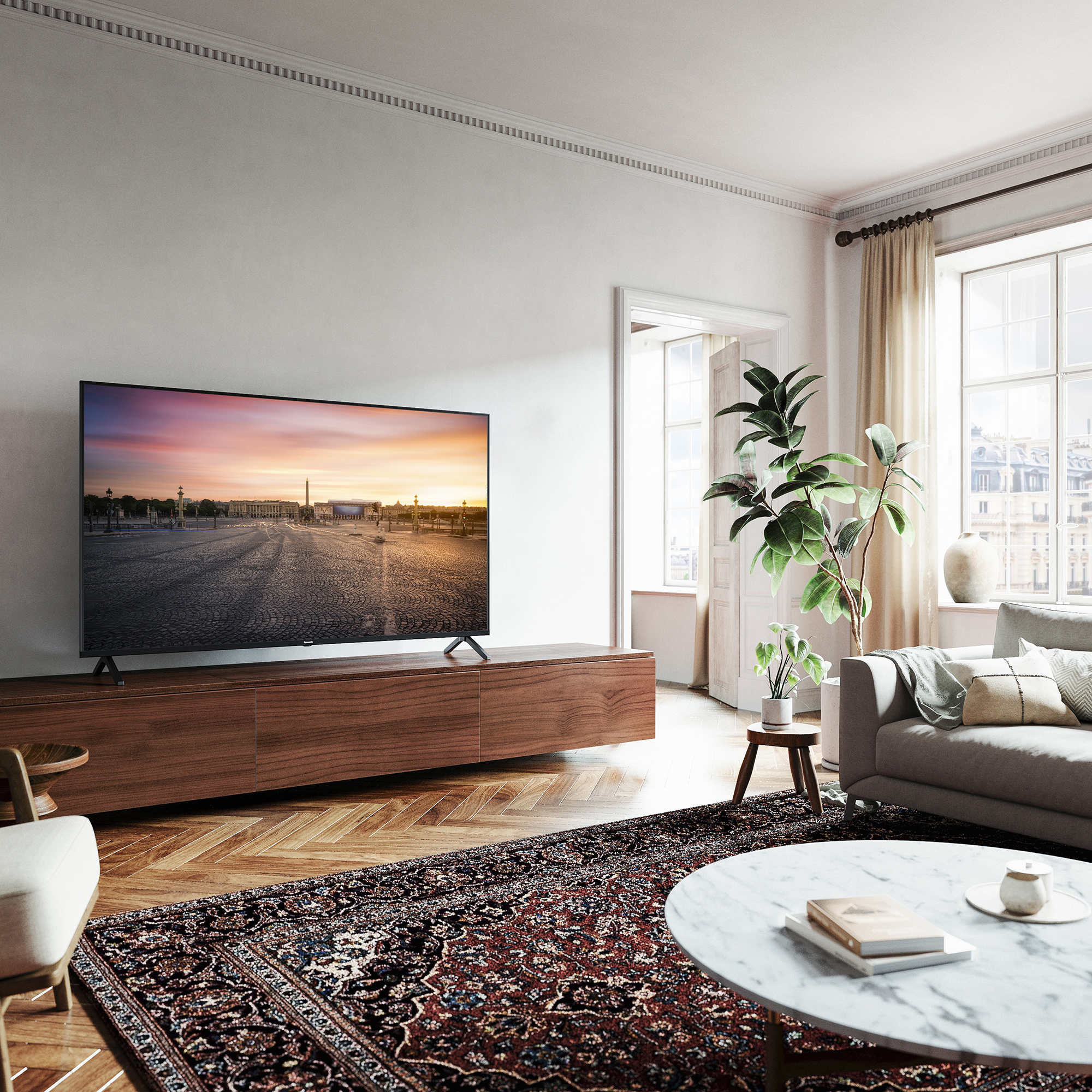 Ein modernes Wohnzimmer mit einem großen Fernseher, der eine Sonnenuntergangs-Stadtszene zeigt, einem grauen Sofa und einem gemusterten Teppich.