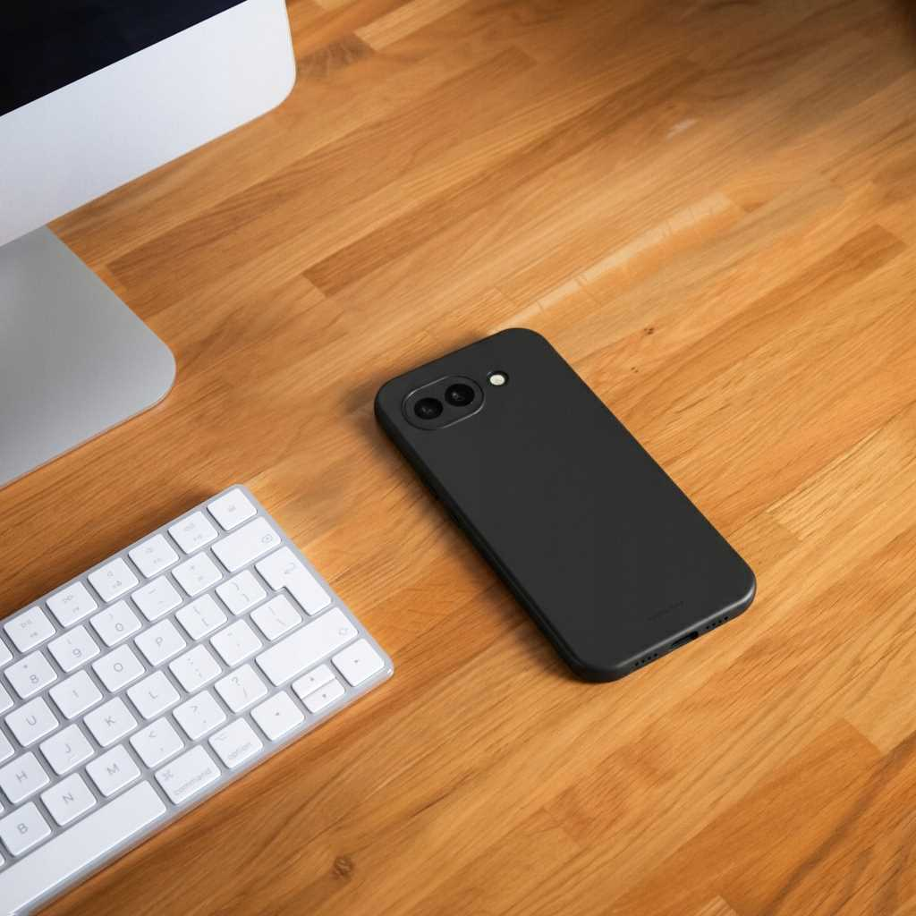 Czarny smartfon leży na drewnianym biurku obok klawiatury i monitora.